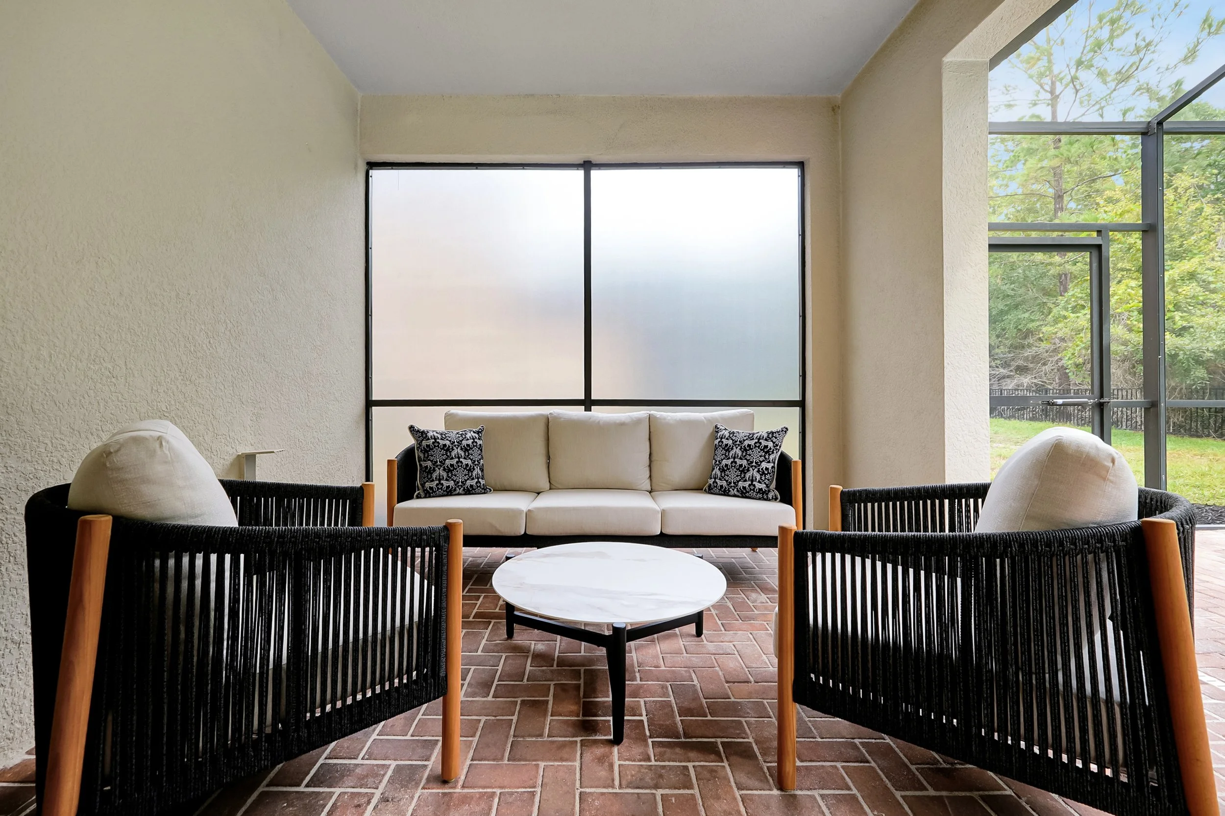 A bright indoor seating area with a white couch, two black and white patterned pillows, black chairs with wooden arms, a round white coffee table, large frosted window, and glass door leading outside with greenery.