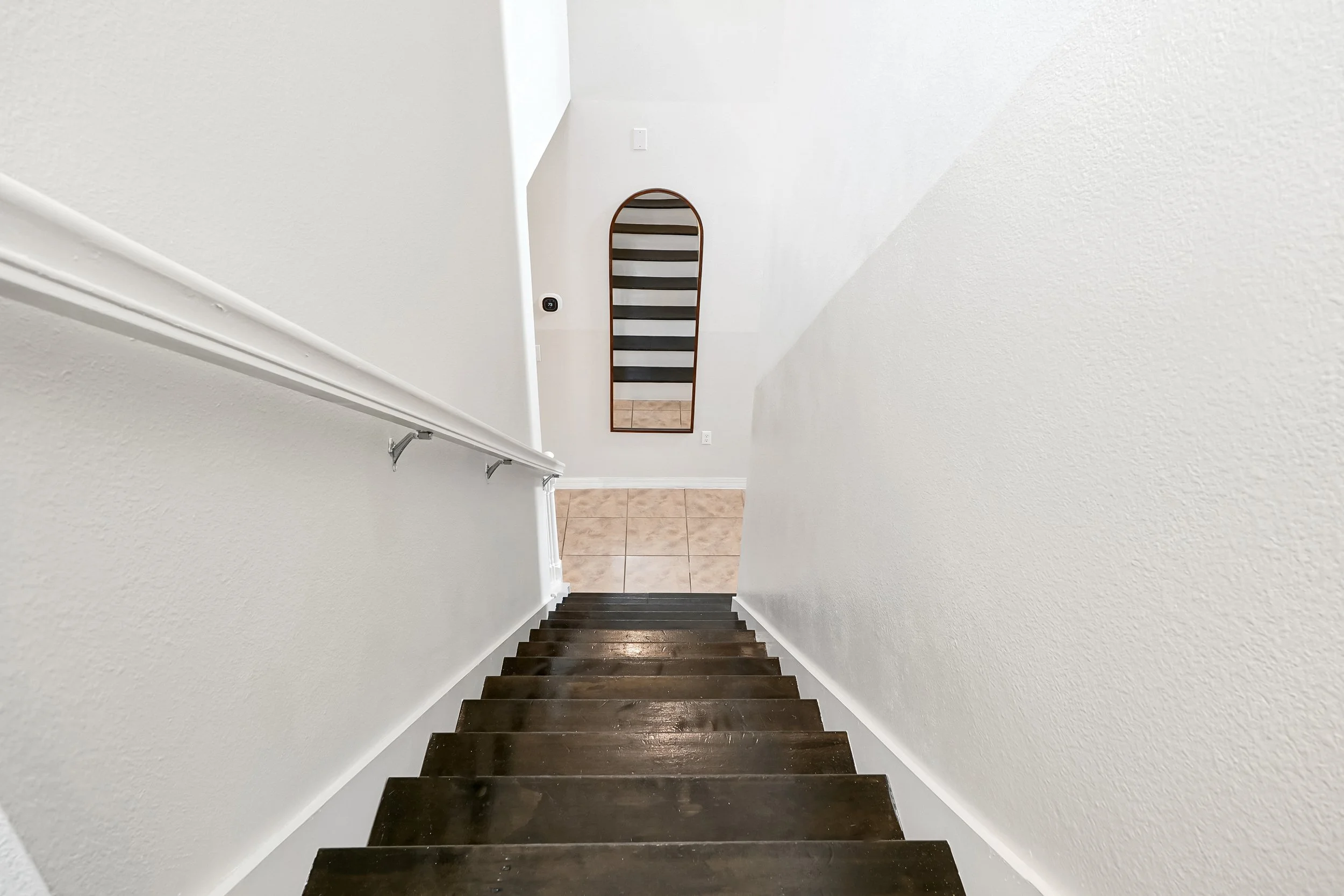 View down a staircase with dark wooden steps, white walls, a handrail on the left, a mirror at the bottom, and tiled flooring beyond the stairs.