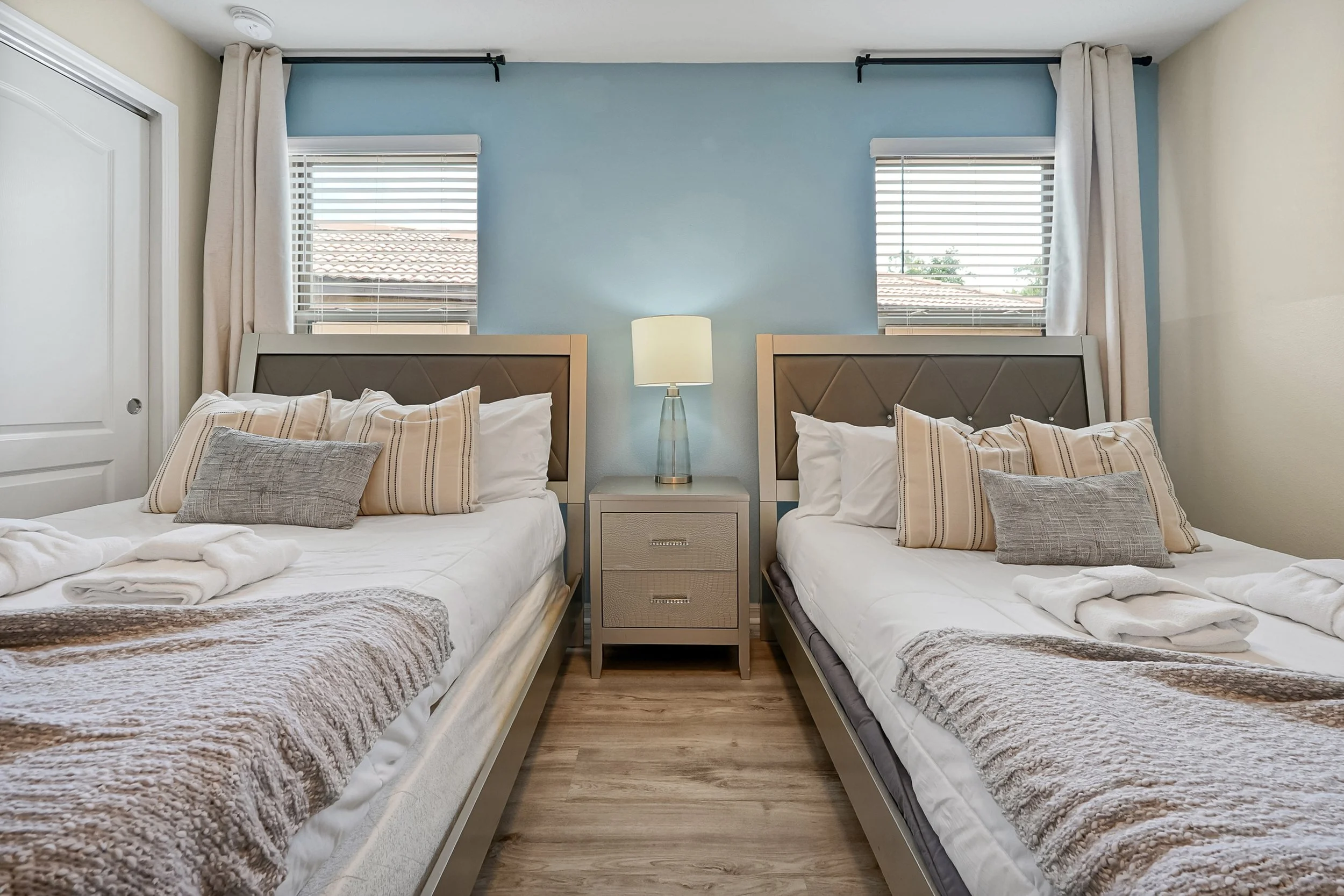 A hotel room with two neatly made beds with beige and white bedding, a wooden nightstand with a table lamp between them, and two windows with white blinds and beige curtains, all with a blue accent wall.