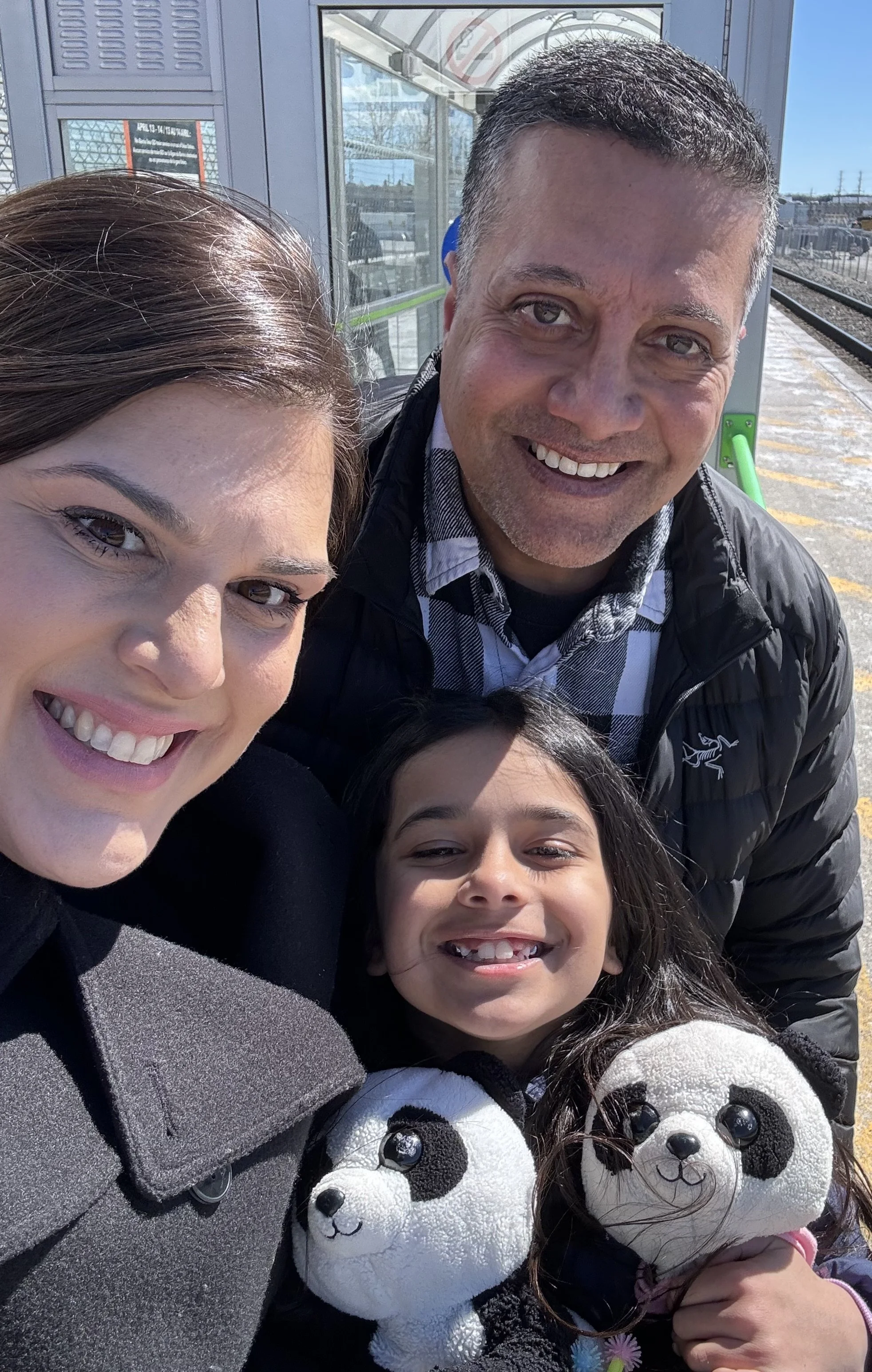 A group of five people taking a selfie outdoors on a sunny day at a train station platform, with three young girls, a woman, and a man smiling and leaning in close together, holding stuffed panda bear toys.