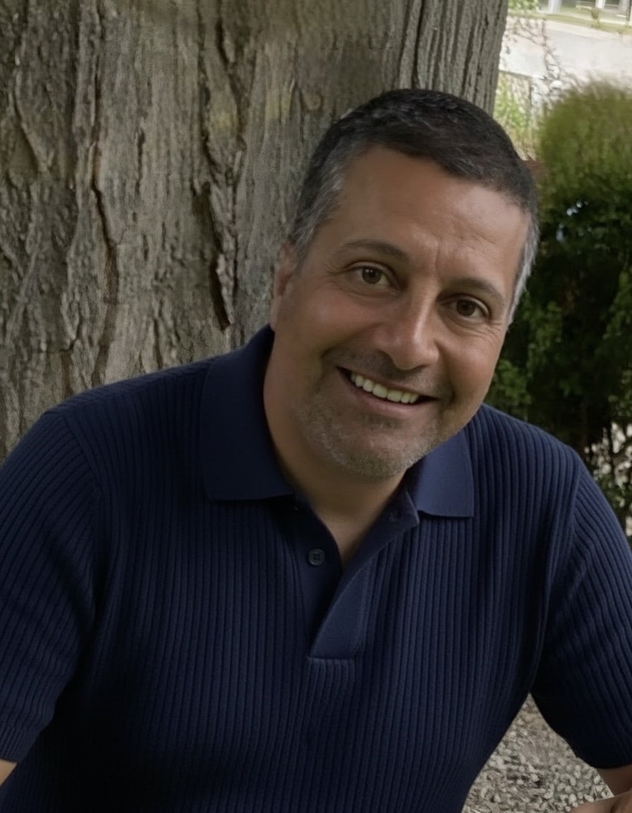 Smiling man leaning against a tree outdoors, wearing a navy blue polo shirt.
