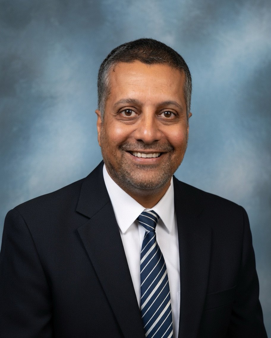 Professional headshot of a smiling man in a dark suit, white shirt, and striped tie against a blue gradient background.