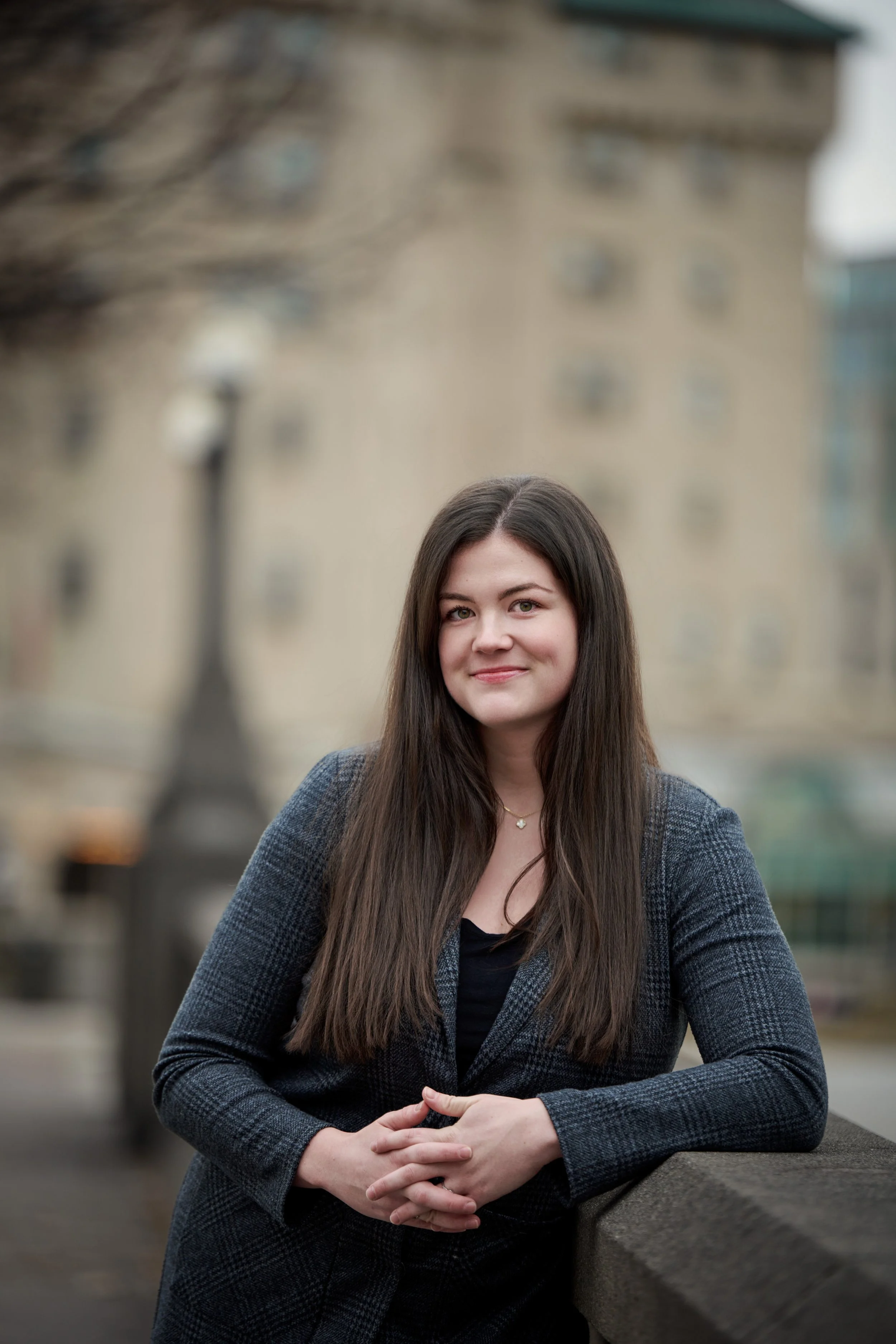 A woman with long brown hair wearing a gray plaid blazer and black top, leaning on a stone railing outdoors, smiling at the camera with a blurred stone building in the background.