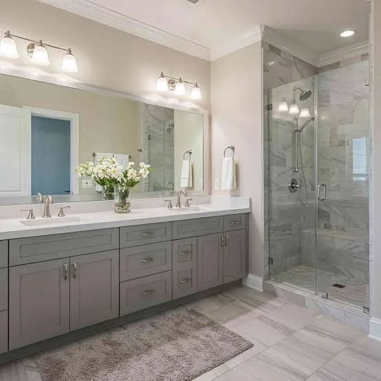 Modern bathroom with gray vanity, large mirror, white countertops, glass-enclosed shower with marble tile, and a vase of white flowers on the counter.