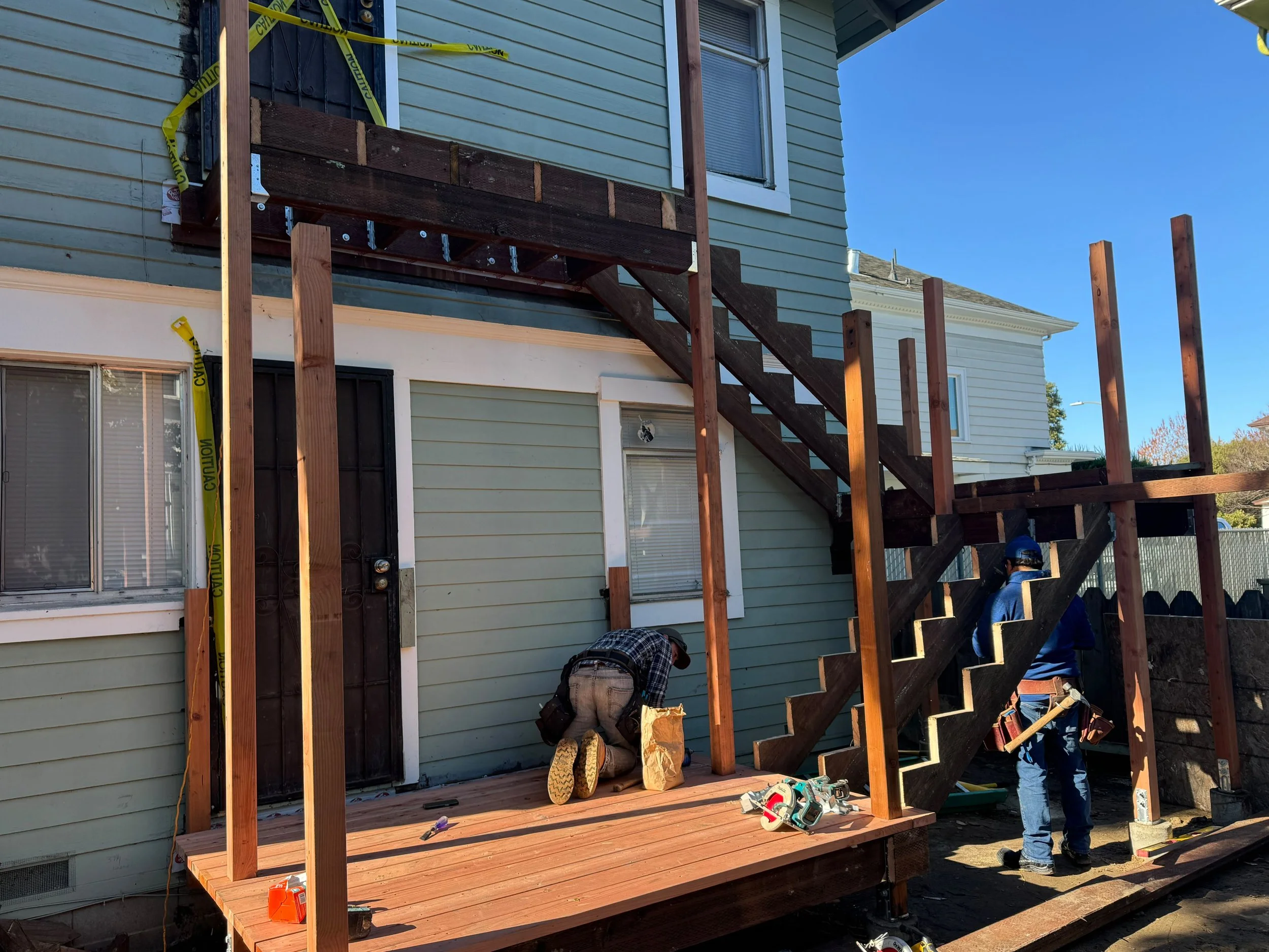 Construction workers building a wooden deck and staircase on the back of a green house, with some yellow caution tape around the area.