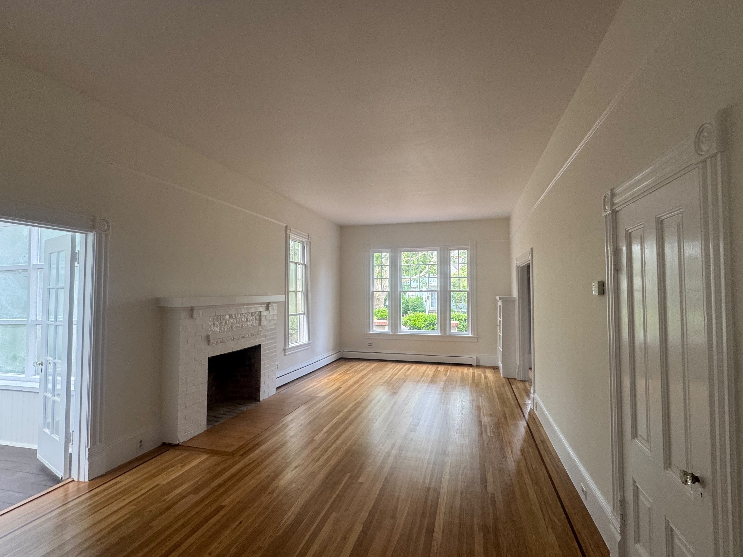 Empty living room with white walls, large windows, hardwood flooring, a fireplace, and a sliding glass door.