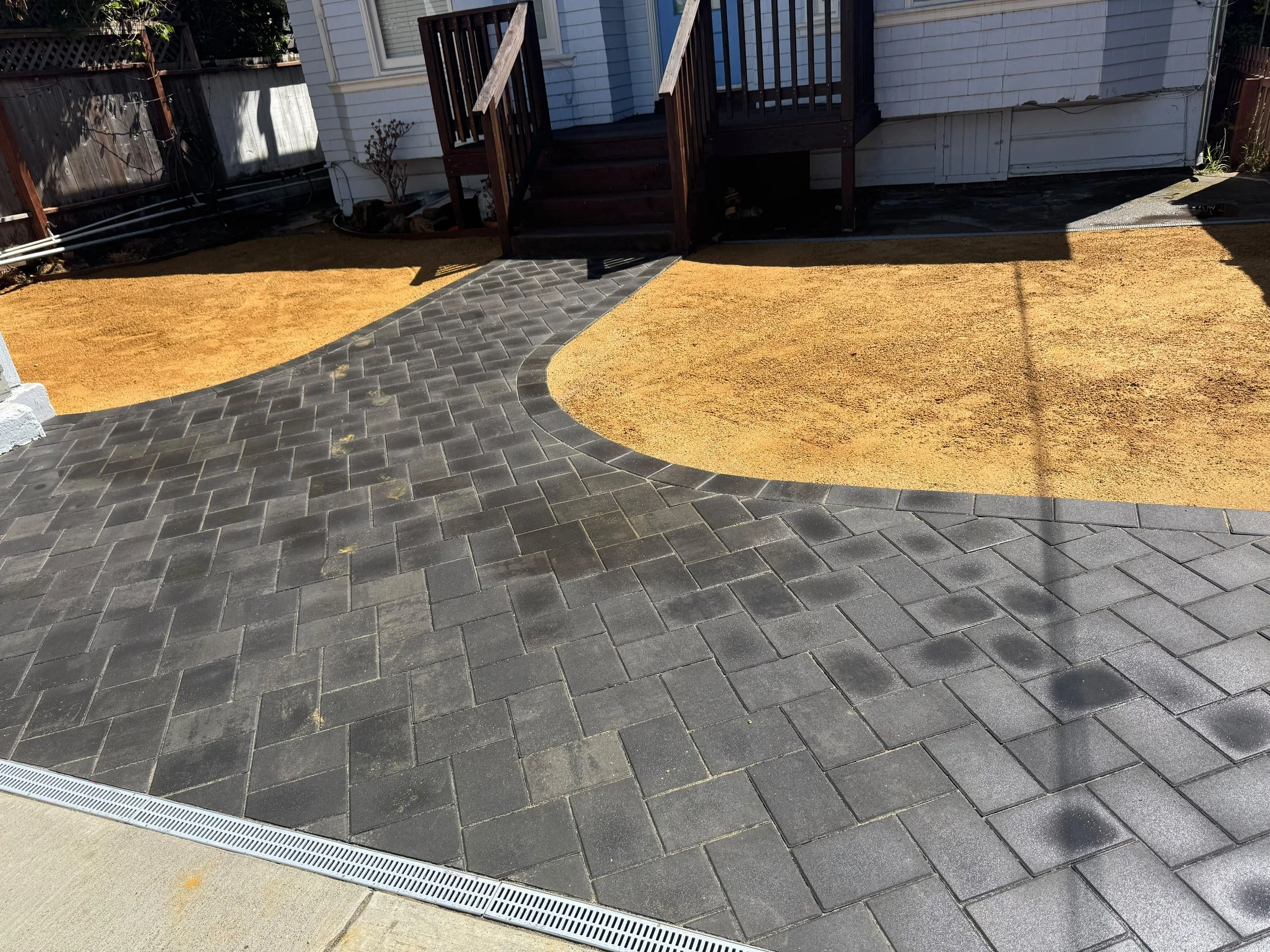 Backyard with a small set of wooden stairs leading to a door, a border of yellow gravel or sand, black paver walkway, and a drainage grate at the edge of the pavers.