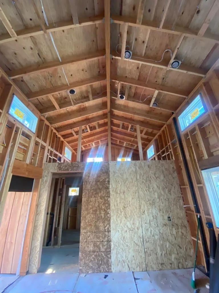 Interior of a house under construction with wooden framing, OSB panels forming a wall, and a high vaulted ceiling with exposed wooden beams and no finished ceiling.