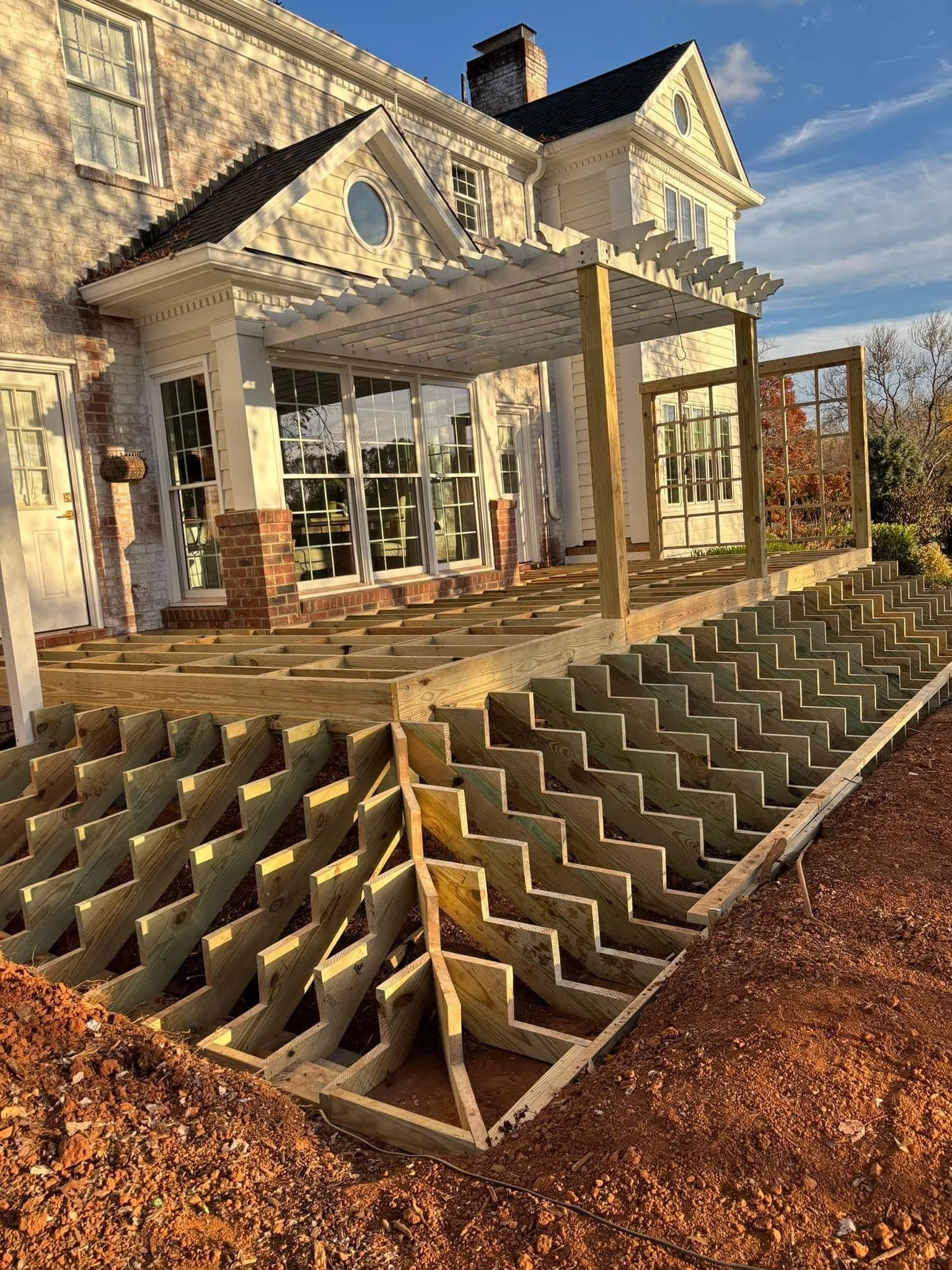 Construction of a wooden deck attached to a house with large windows and brick columns, with the decking framework in progress on a dirt ground.