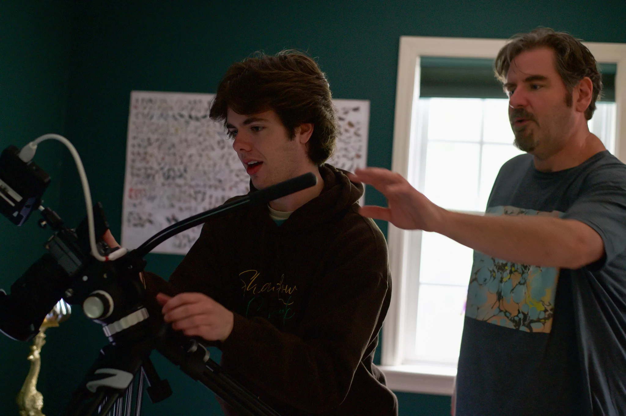Two men looking at a camera mounted on a tripod in a room with dark green walls and a window. One man is young with dark hair, the other has gray hair and beard.