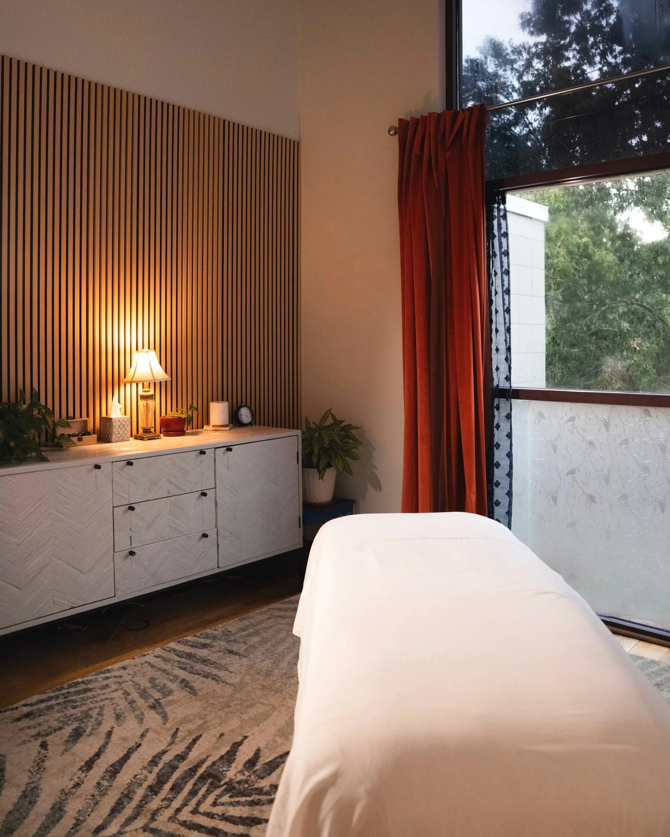 Inviting room interior with a massage table in the foreground, a white dresser with a lamp, a window with subtle orange curtains, and a view of trees outside.