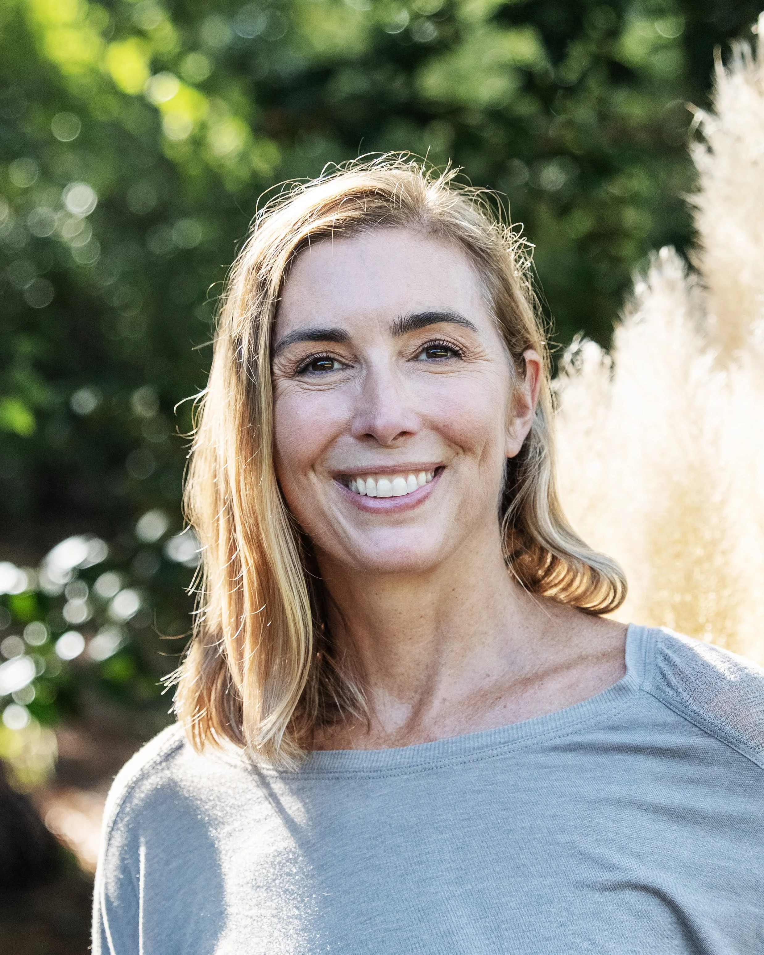 Close-up of a smiling woman with blonde hair outdoors in natural sunlight, wearing a light gray top.
