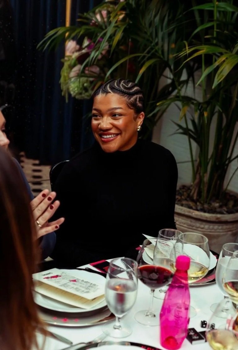 A woman with a short, braided hairstyle smiling and engaged in conversation at a dinner table, with wine glasses and a pink water bottle on the table, surrounded by greenery.