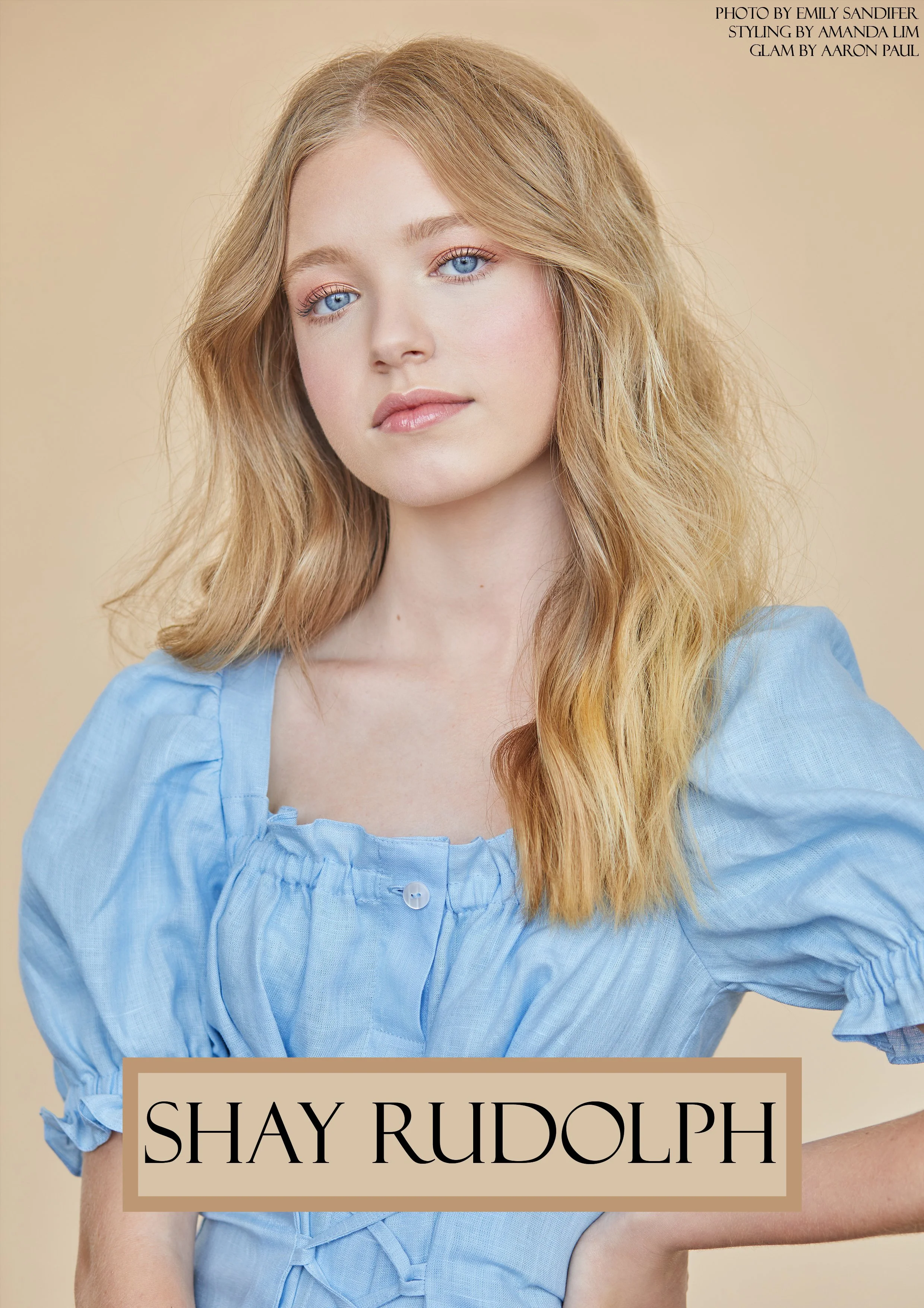 A woman with long, wavy blonde hair and blue eyes wearing a light blue blouse, against a beige background. The text on the image credits Emily Sandifer for the photo, Amanda Lim for styling, and Aaron Paul for glam.