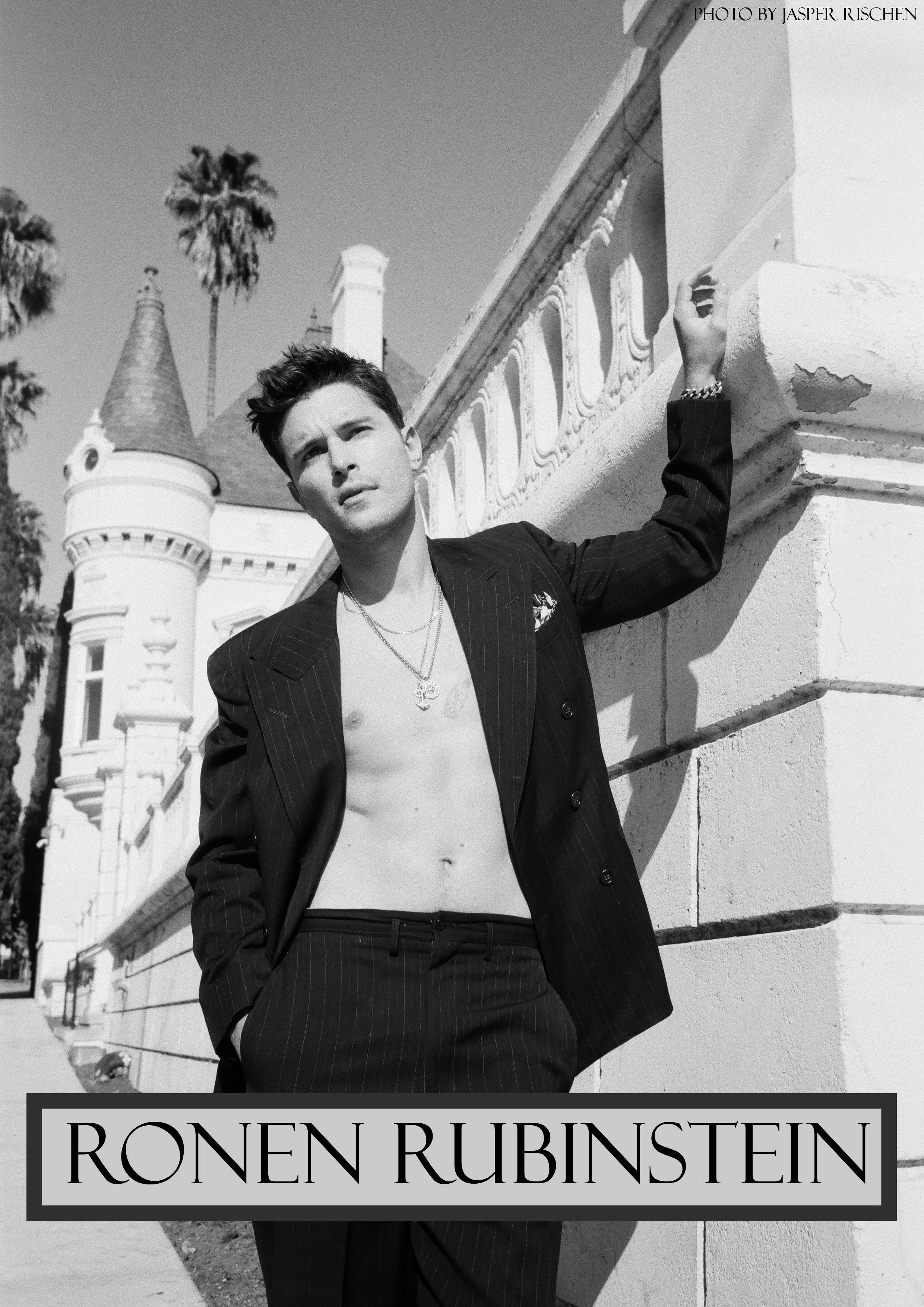 Black and white photo of a young man with dark hair, dressed in a pinstripe suit jacket with no shirt underneath, leaning against a concrete wall with ornate detailing. Tall palm trees and a castle-like building with turrets are in the background. Text on the image reads "RONEN RUBINSTEIN" and "PHOTO BY JASPER RISCHEN."