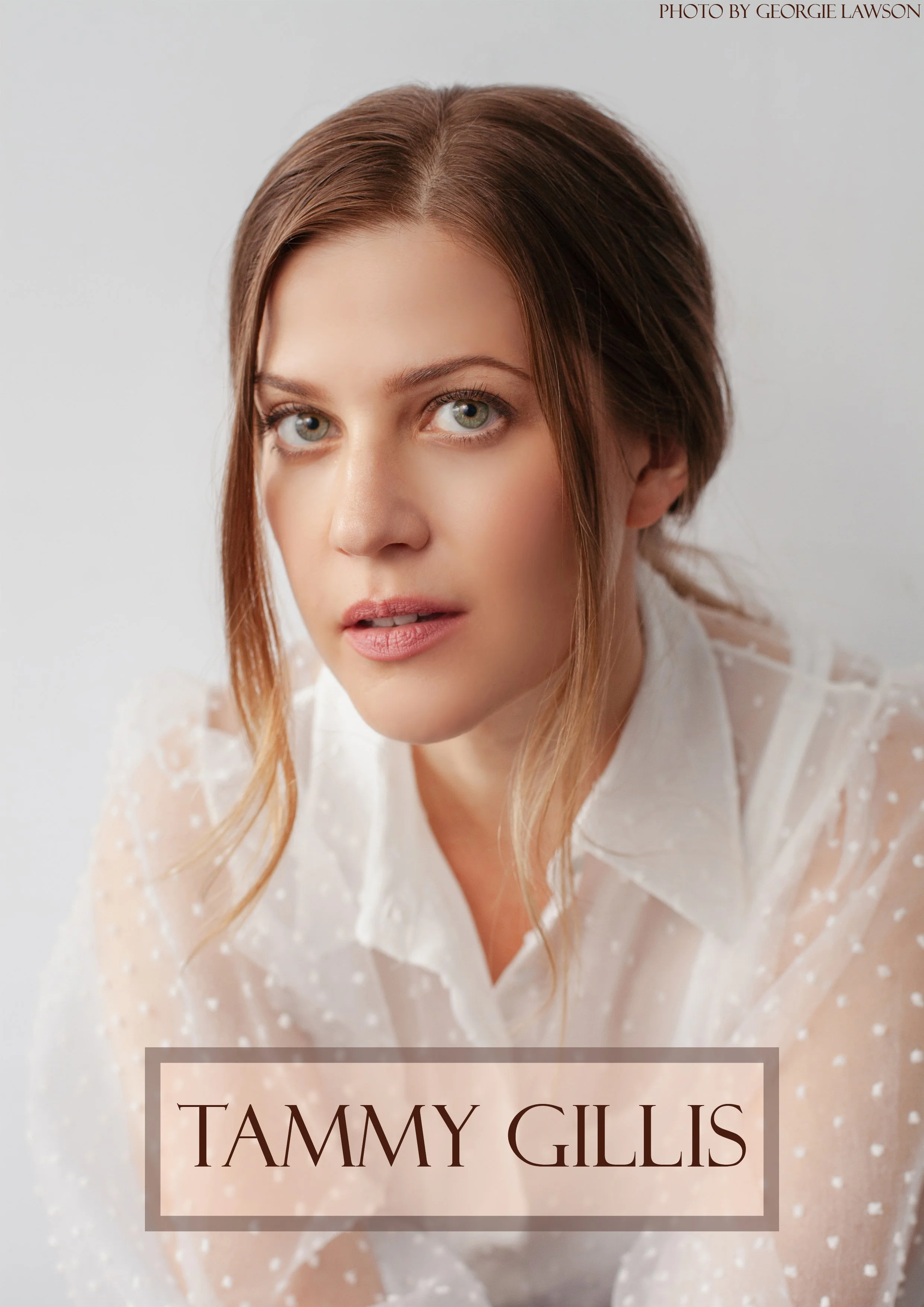 A woman with light skin, blue eyes, and long red hair styled in loose waves, wearing a white dress with a sheer polka-dot pattern and lace details, looks directly at the camera with a soft expression. The background is plain and light-colored.