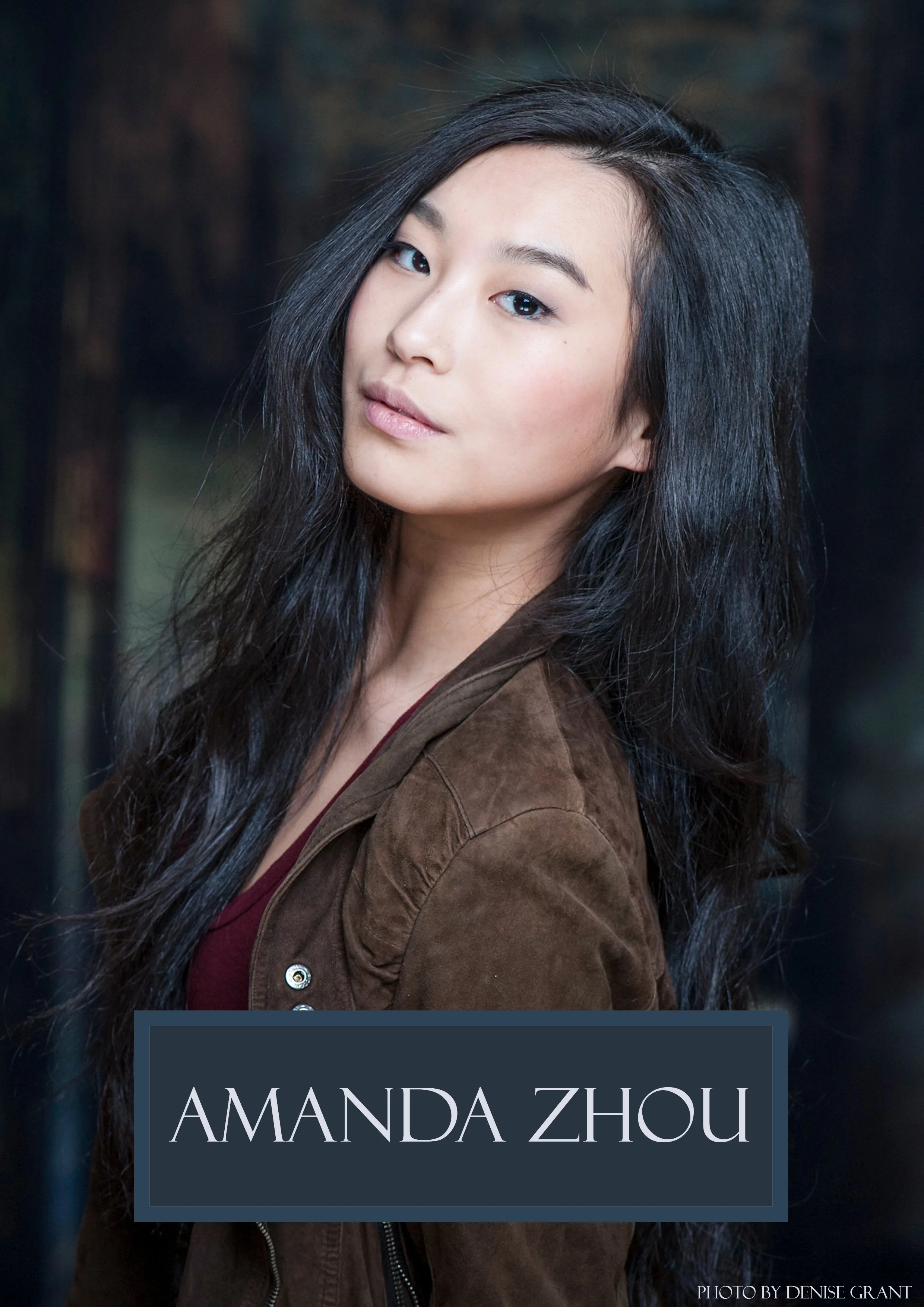 Close-up portrait of a young woman with long black hair, wearing a brown jacket and a dark top, looking at the camera with a slight smile against a dark background. Text overlay: 'Amanda Zhou' and photo credit 'Photo by Denise Grant'.