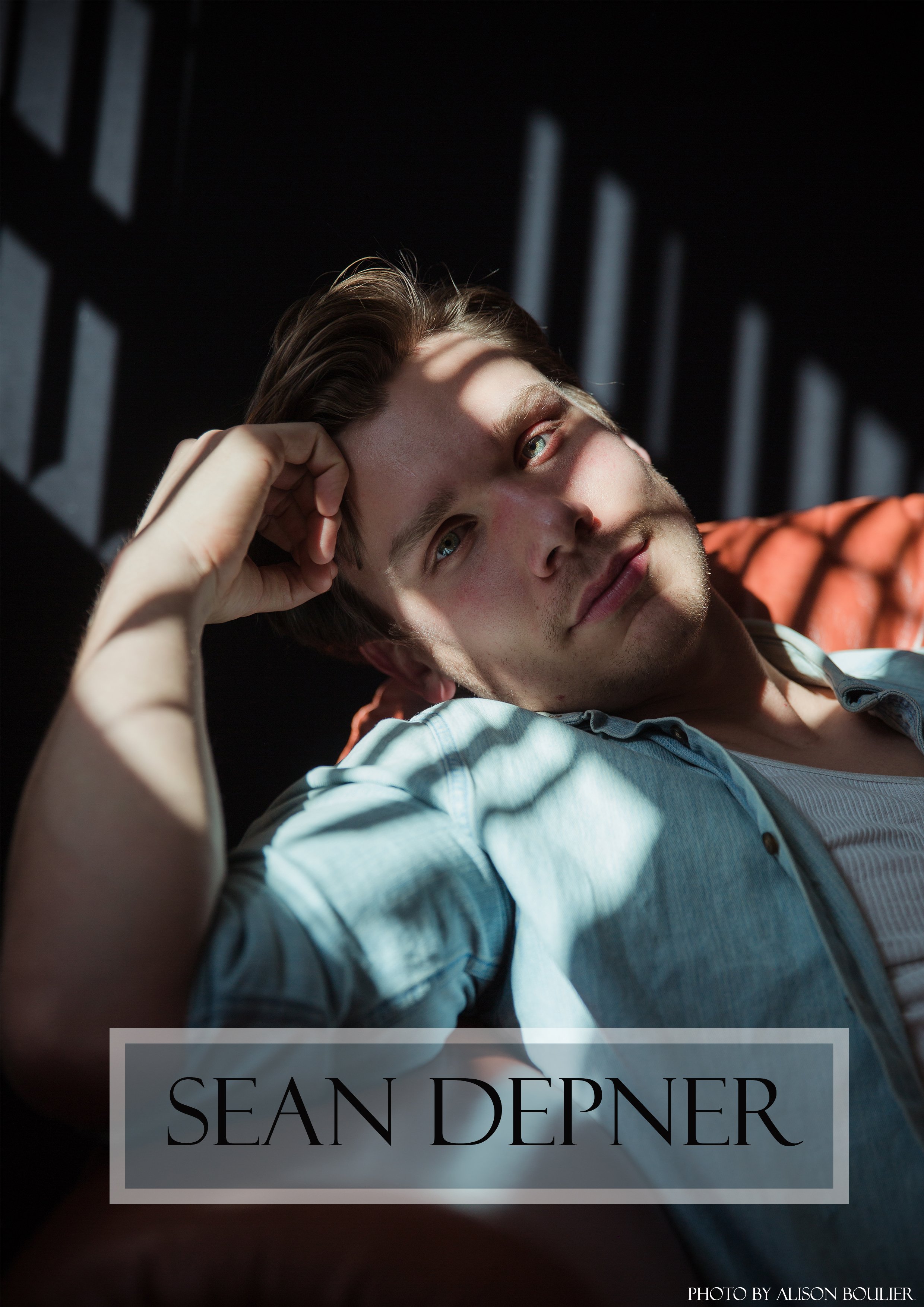 Close-up of a young man with light brown hair and blue eyes lying on a bed, resting his head on his hand, with sunlight and shadows creating patterns on his face and surroundings.