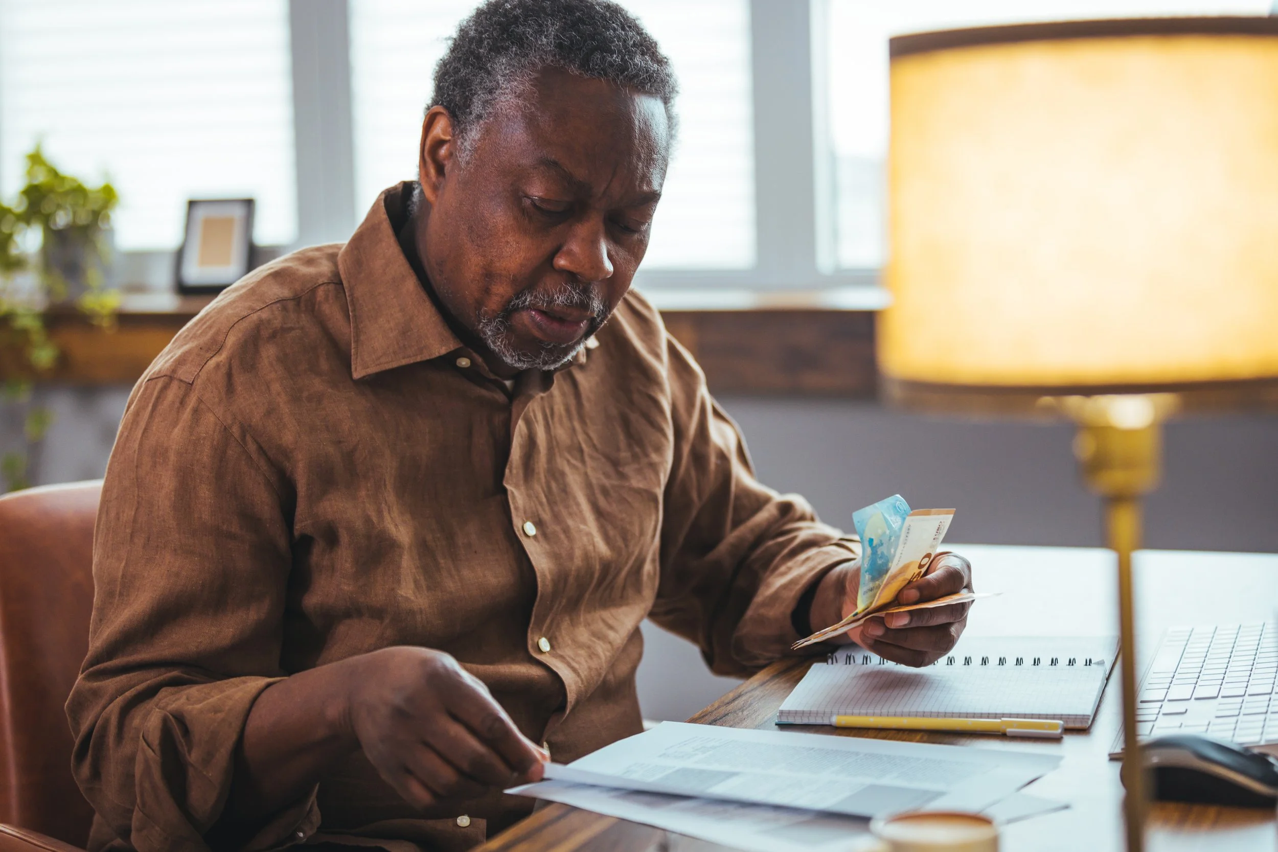 An elderly man with gray hair and facial hair is sitting at a desk, looking at money in his hands. The desk has a notebook, papers, and a ruler on it. There is a table lamp and some framed pictures in the background, with natural light coming through the windows.