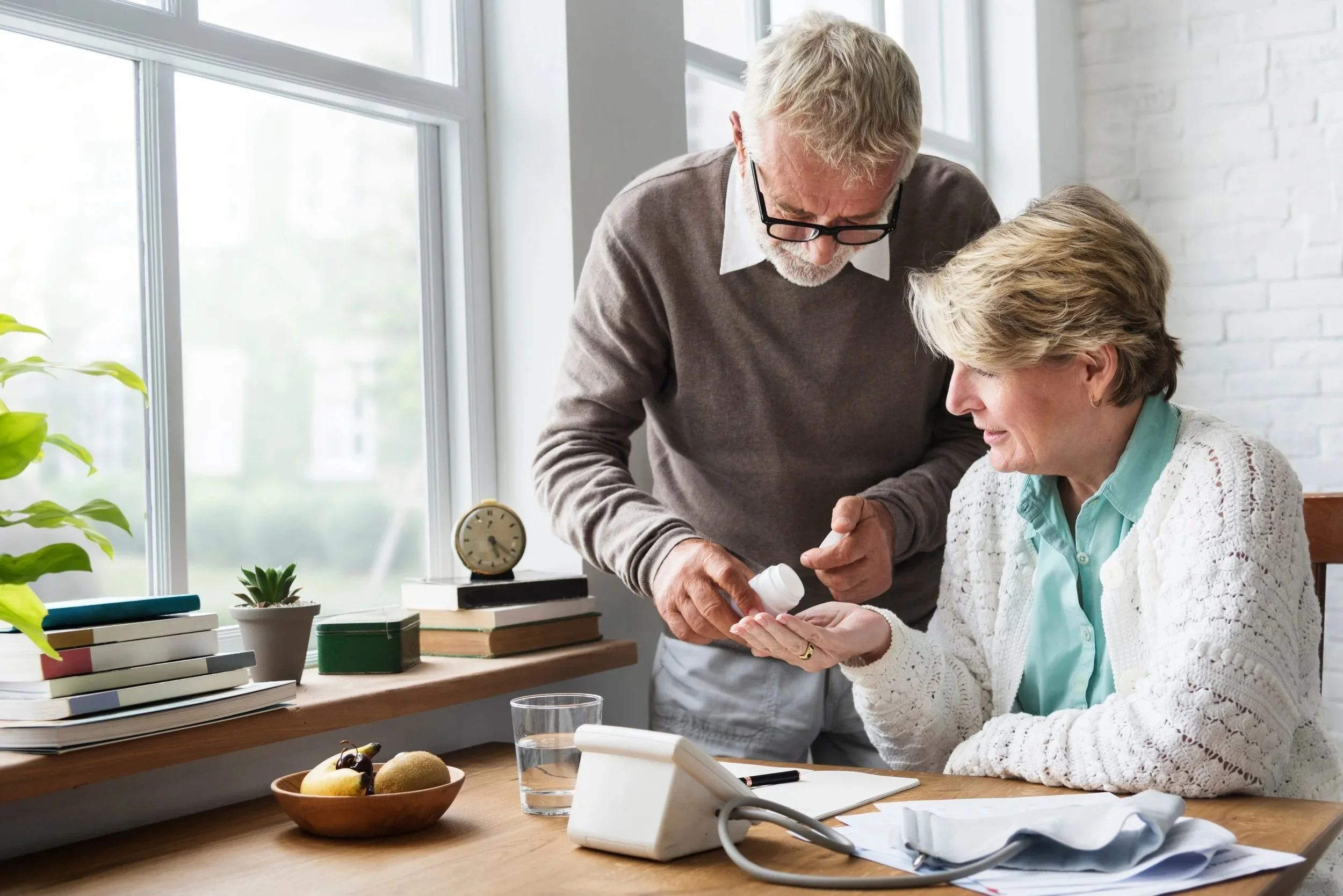 An elderly man and woman are sitting at a desk near a large window. The man is handing something to the woman, possibly medication or a small item. The desk has books, a potted plant, a glass of water, a telephone, and papers. The scene suggests a caring interaction, possibly in a home or care setting.