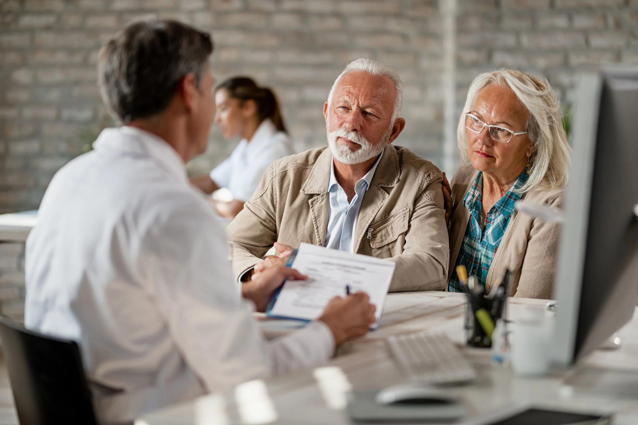 A group of elderly people sitting at a table during a medical consultation or appointment, with a doctor or healthcare professional talking to them.