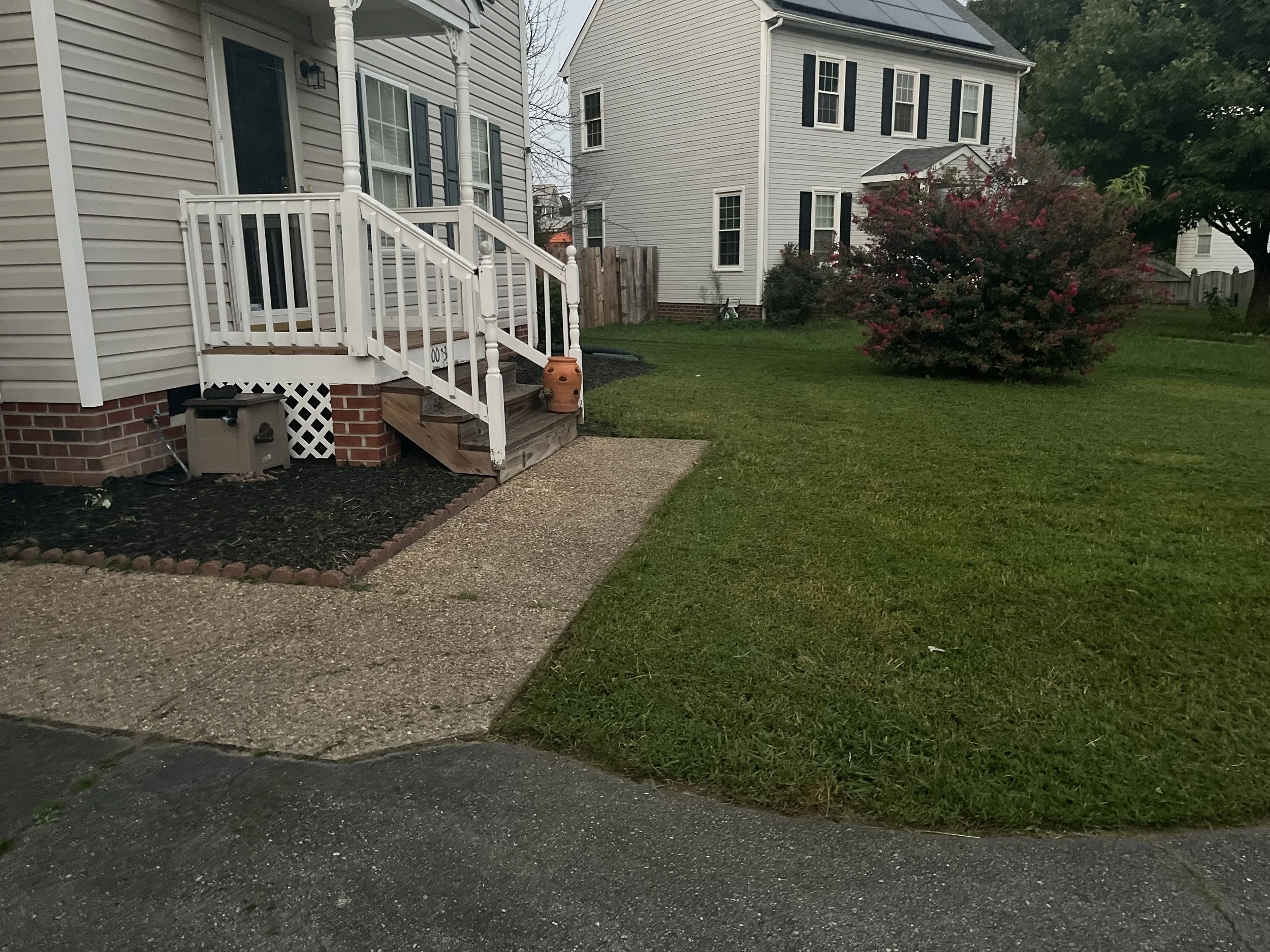 A suburban backyard with a small porch, green lawn, trees, and a fence. The house has beige siding and black shutters.