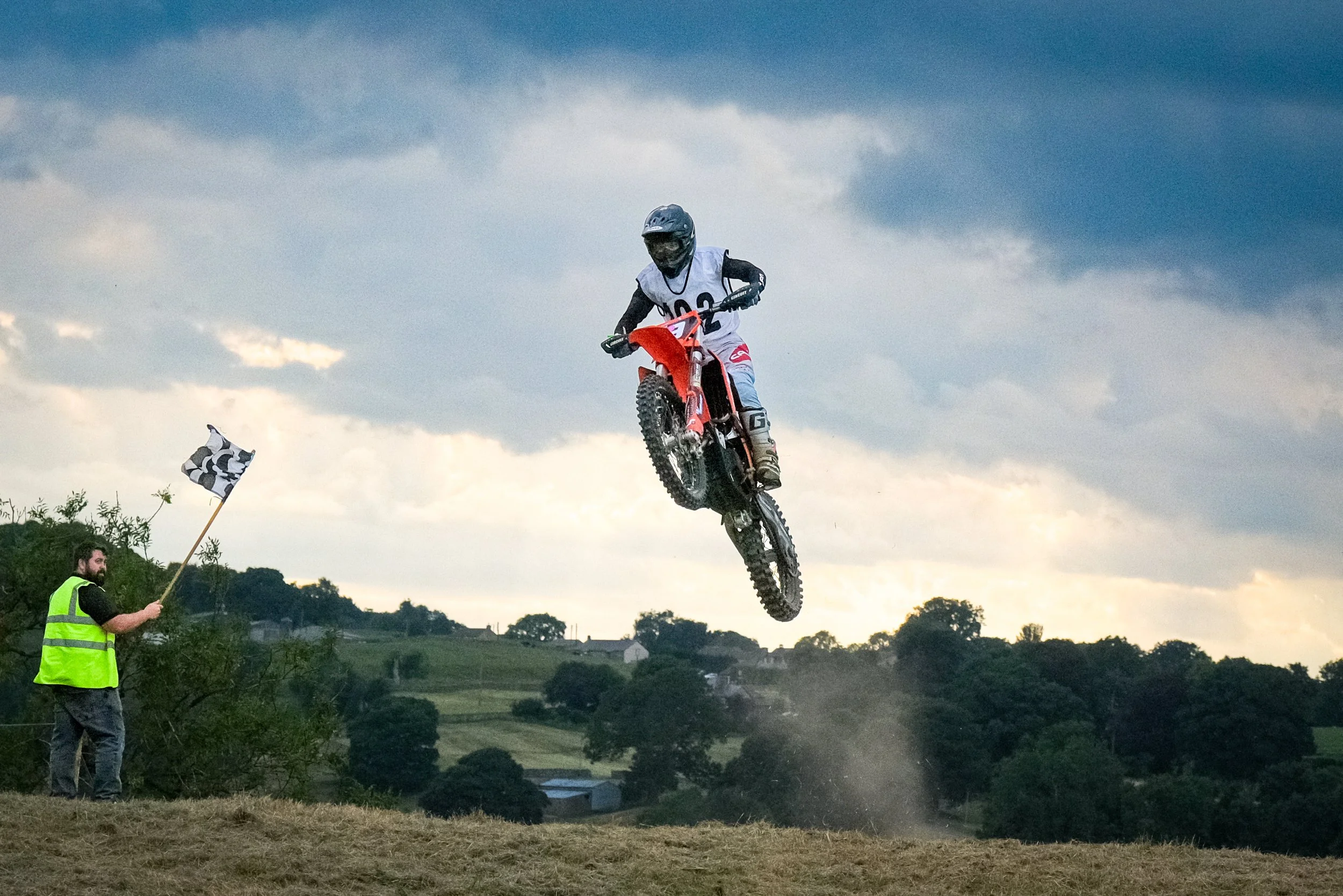 Motocross rider in mid-air jump during race at sunset, with a race official holding a checkered flag in the foreground.