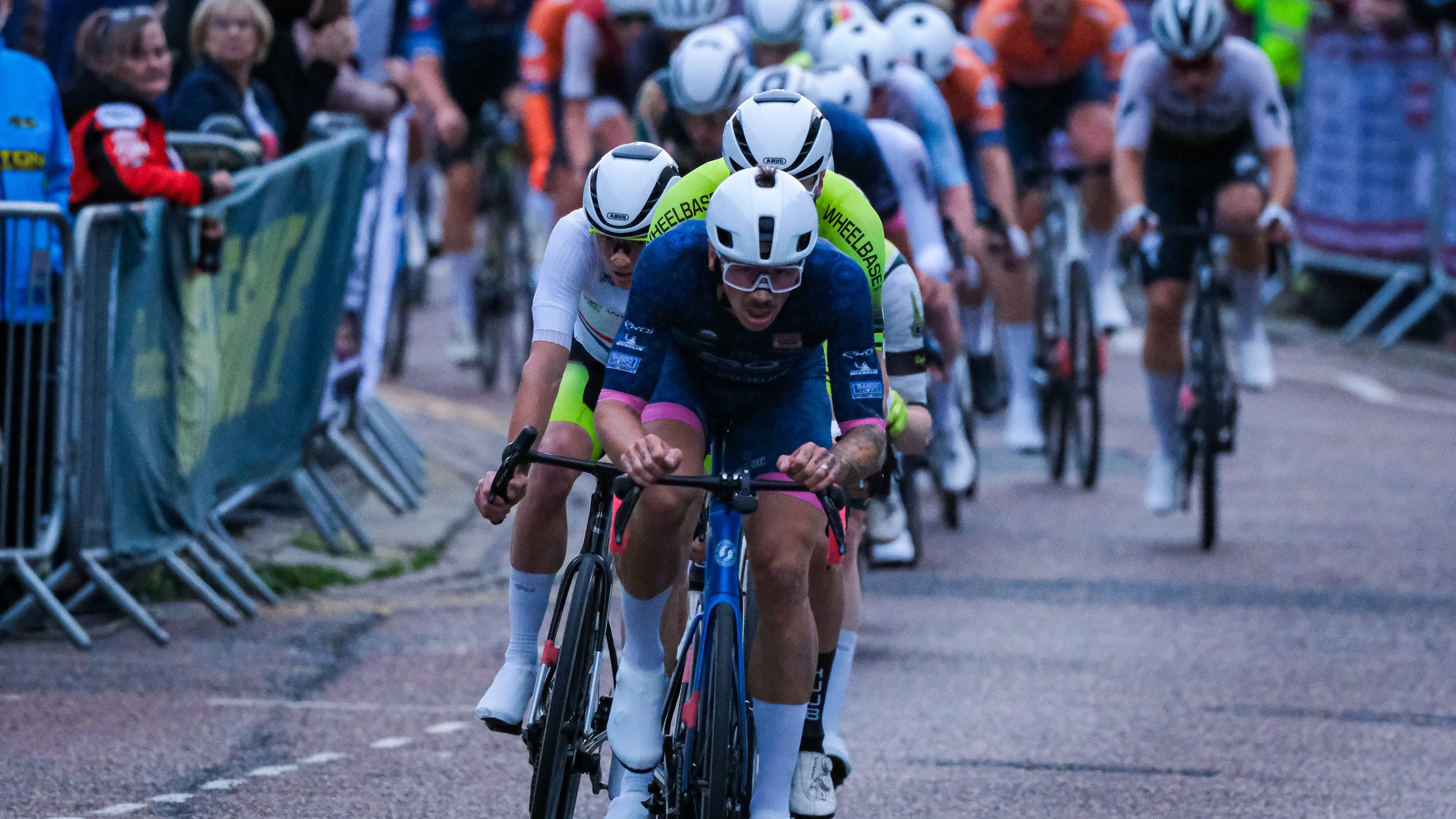 Cyclists competing in a race, wearing helmets and athletic gear, surrounded by spectators behind barriers.