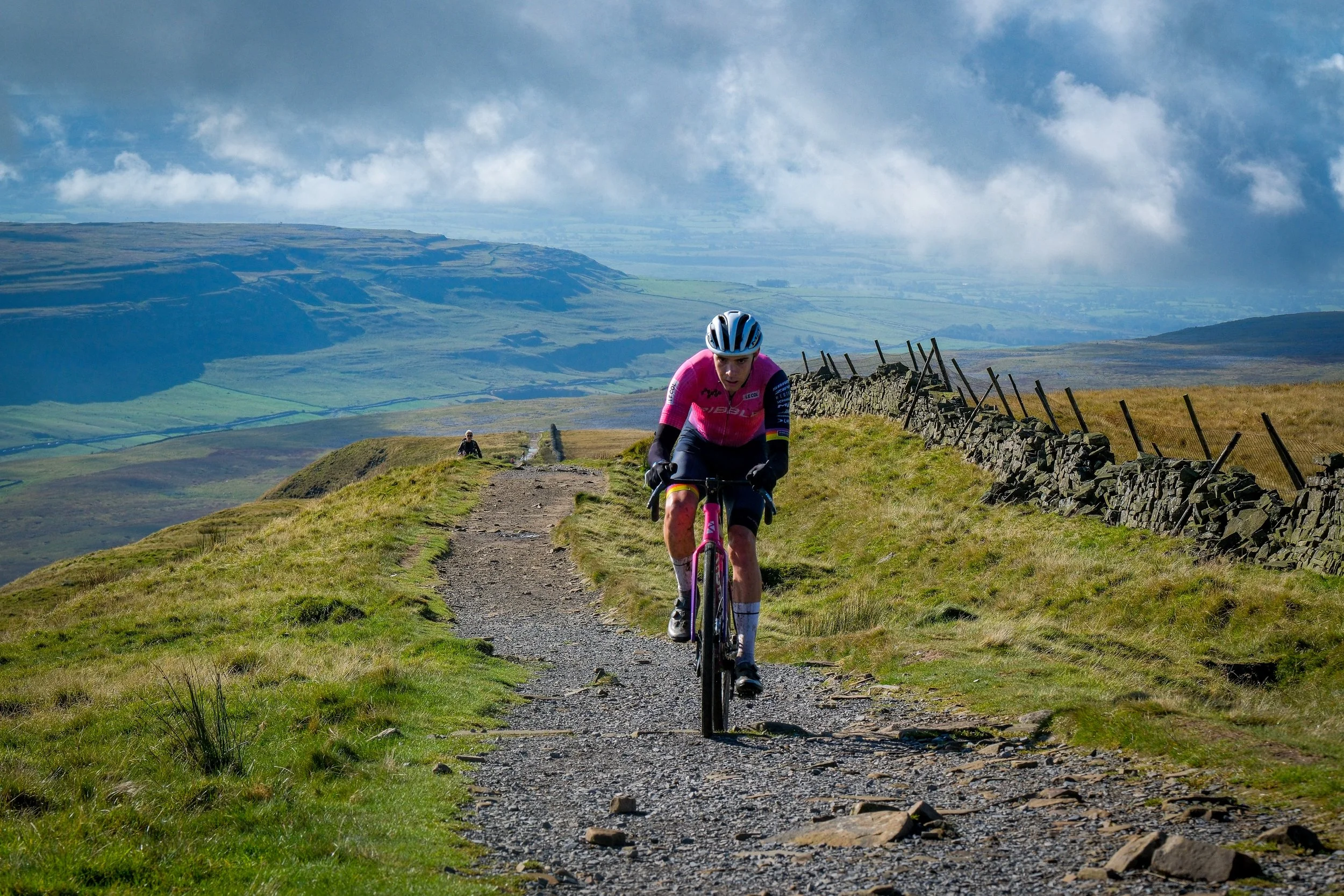 A person wearing a pink cycling jersey and helmet riding a mountain bike on a rugged dirt path in a hilly landscape with green fields, stone fences, and a cloudy sky.