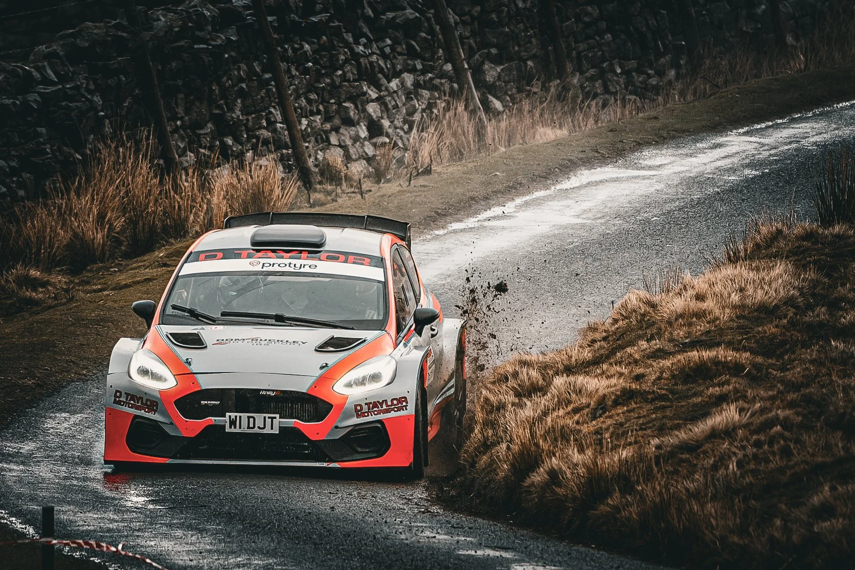 A rally car driving on a wet, narrow rural road with grass on the sides and a stone wall in the background.