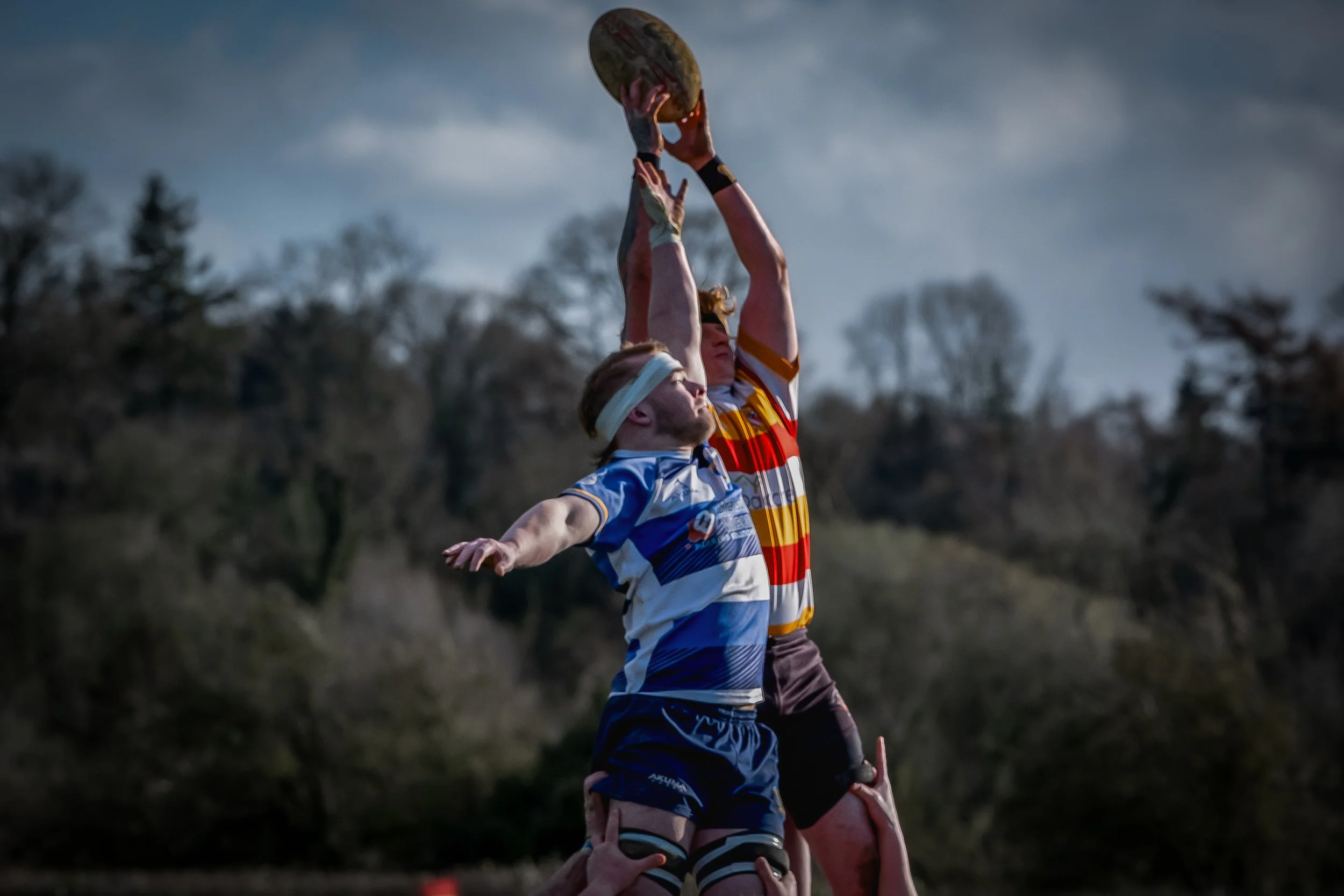 Two rugby players, one in a white and blue jersey and the other in a red and yellow striped jersey, reach upward to catch a rugby ball against an outdoor background with trees and a cloudy sky.