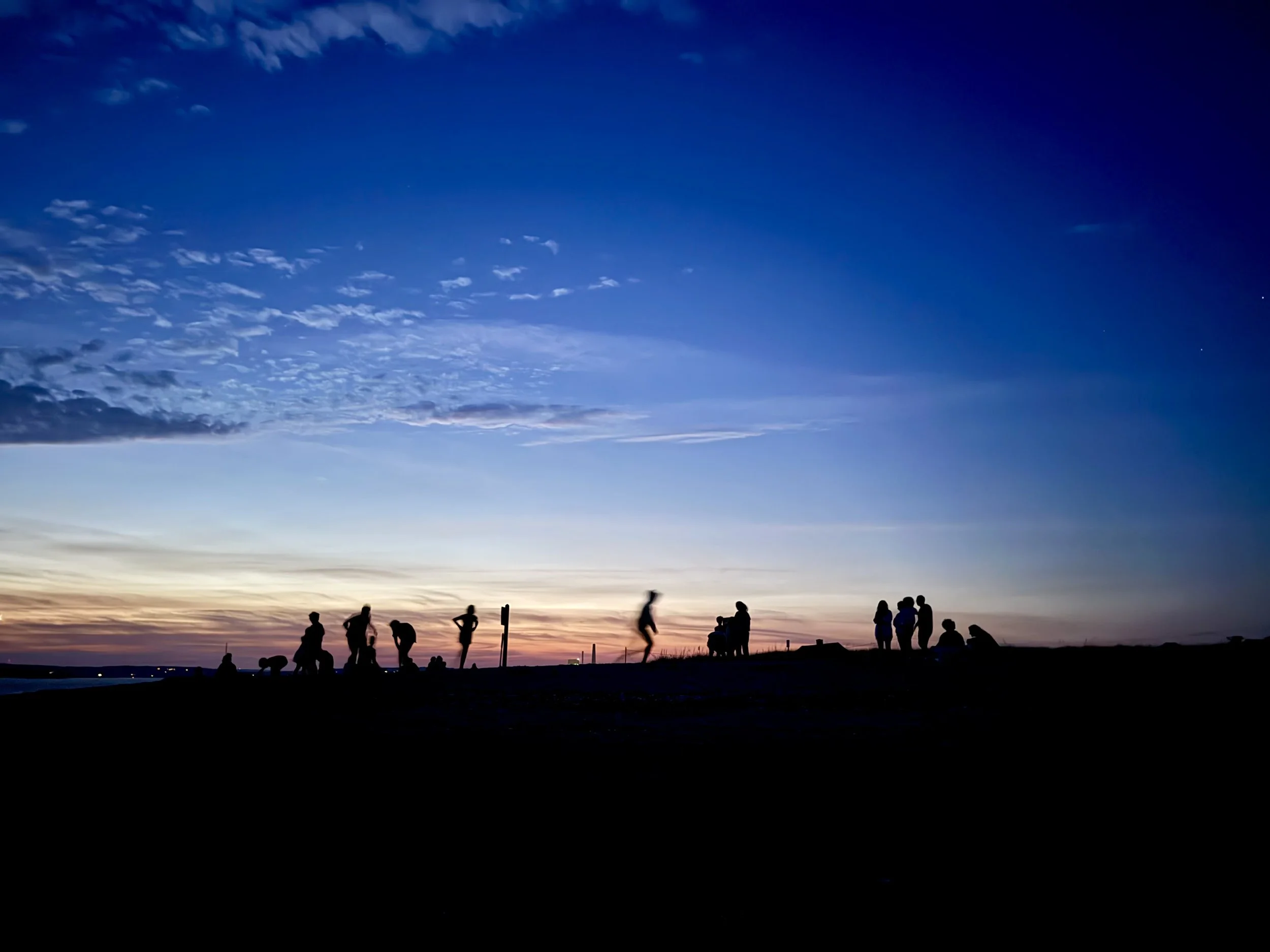 Silhouettes of people sitting and walking along Indian Point beach during sunset, with a sky transitioning from orange near the horizon to dark blue at the top, scattered clouds, and a visible star.