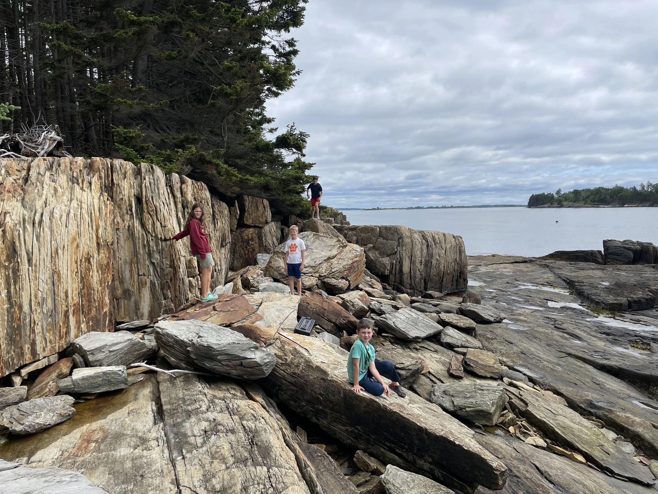 Four kids playing on rocks on Deer Point, Chebeague Island, near a body of water with cliffs and trees in the background under a cloudy sky.