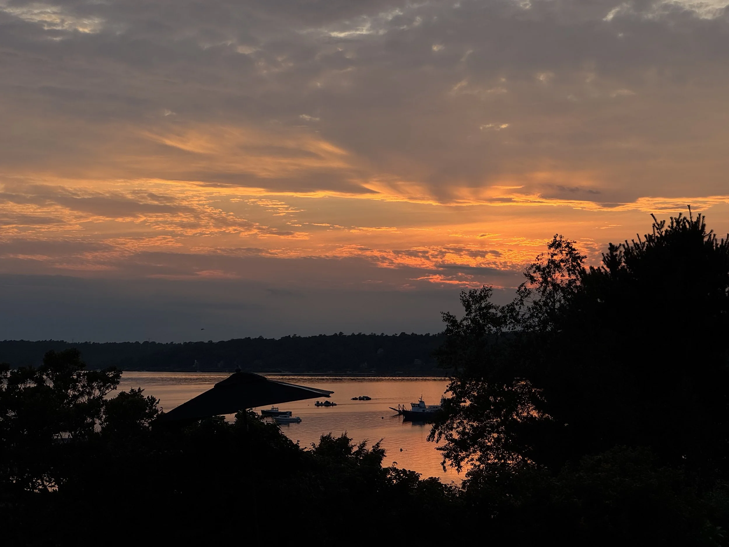 A sunset over Casco Bay, taken from the Chebeague Island Inn, with trees and an umbrella in the foreground.
