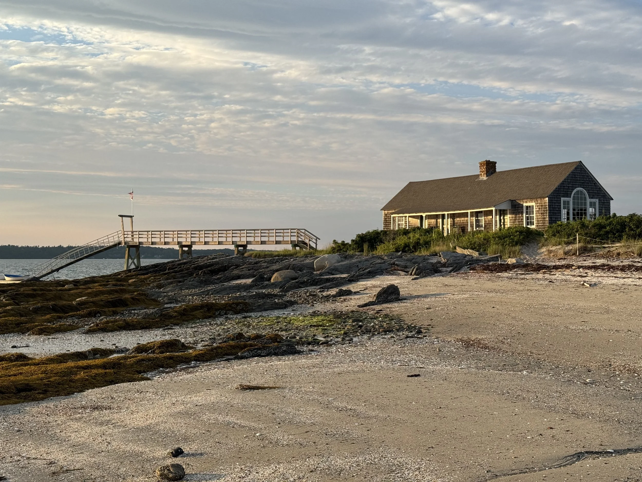 A beach house Indian Point, Chebeague Island Maine, on a grassy hill with large windows and a chimney, next to a pier extending into water with an American flag, under a partly cloudy sky.