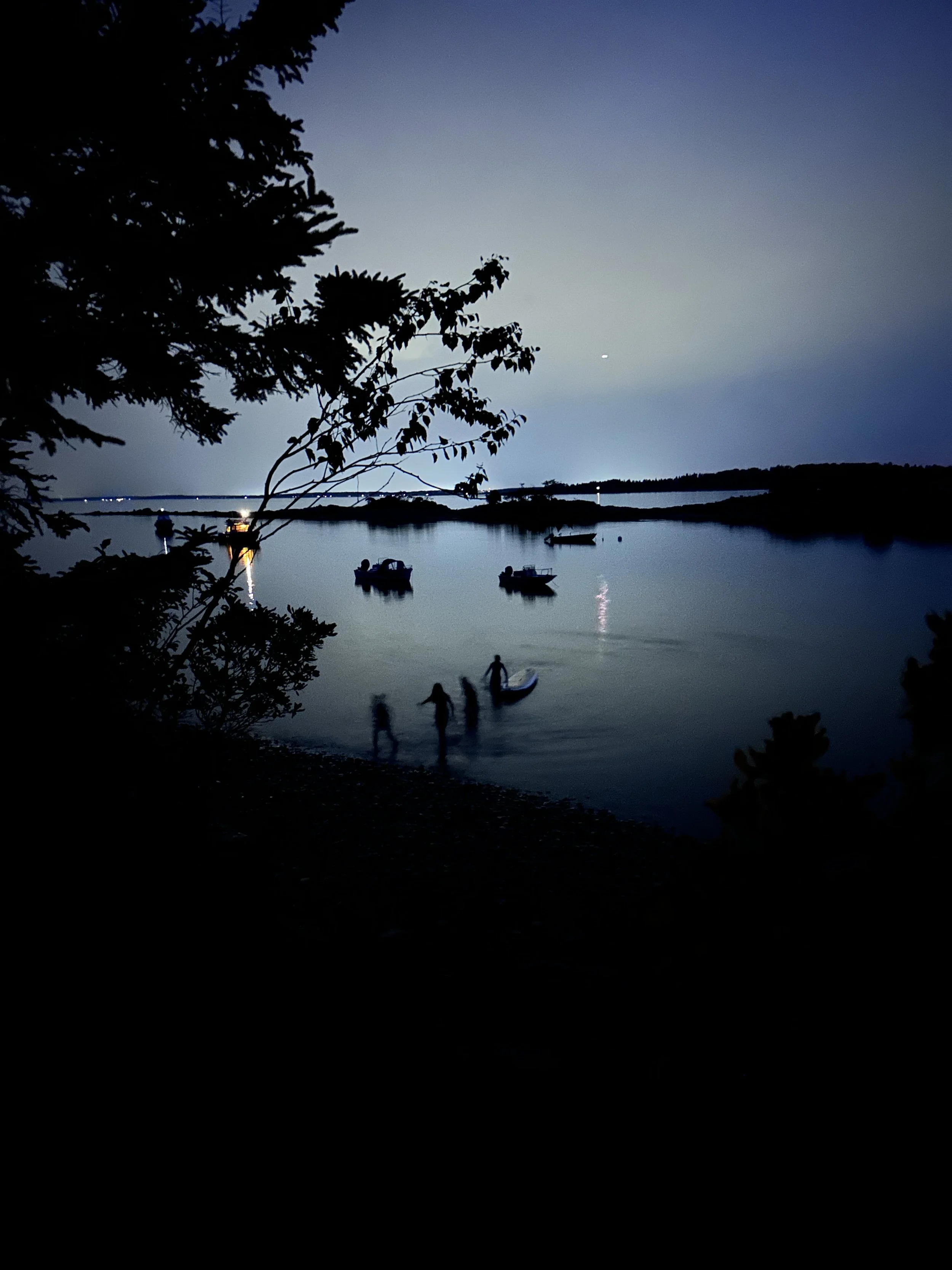 A calm body of water at dusk with boats floating, a person paddleboarding, and trees in the foreground. The sky is a gradient of blue and the scene is peaceful and serene.