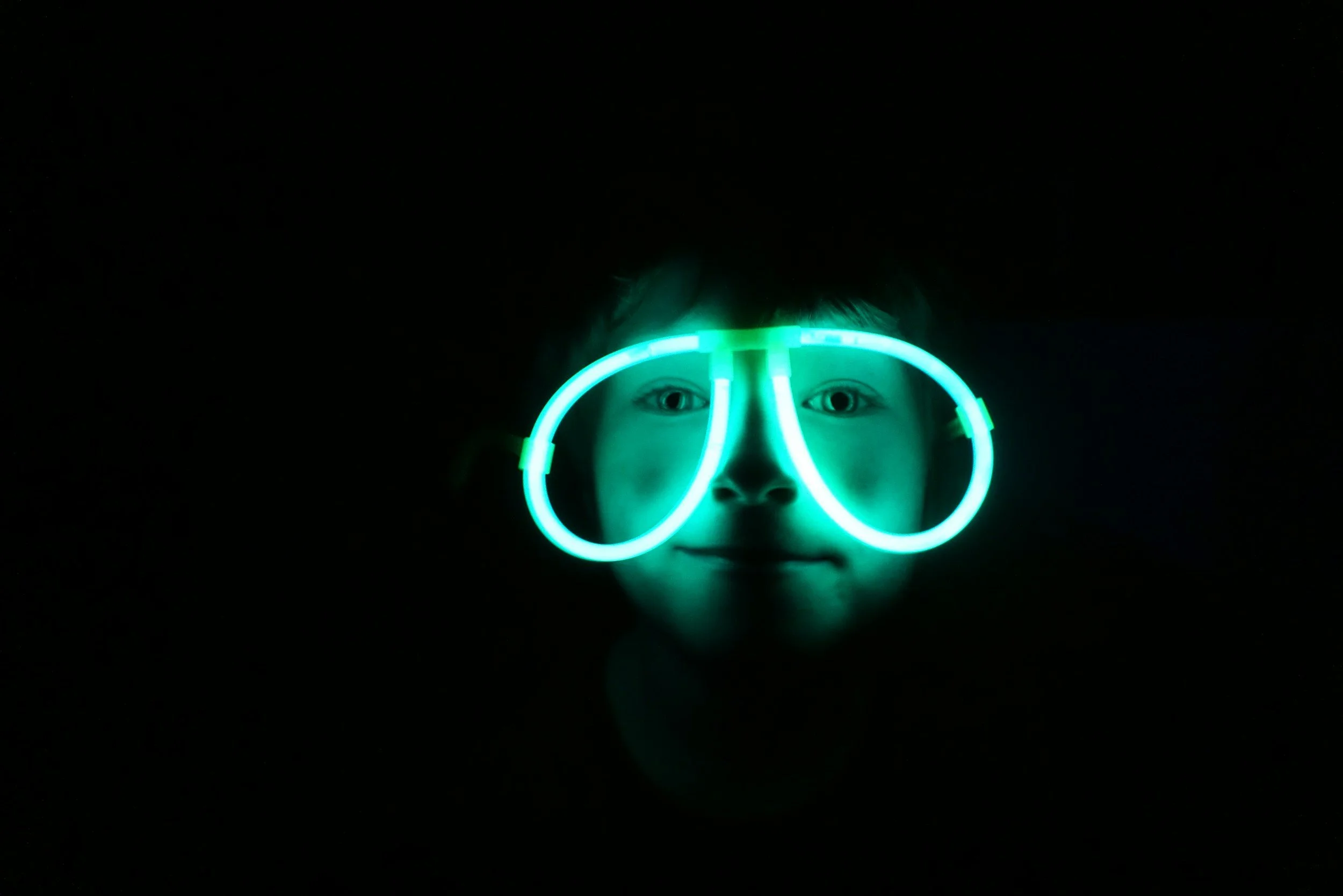 A child’s face is illuminated by glowing neon glasses, creating a luminous effect in the darkness.