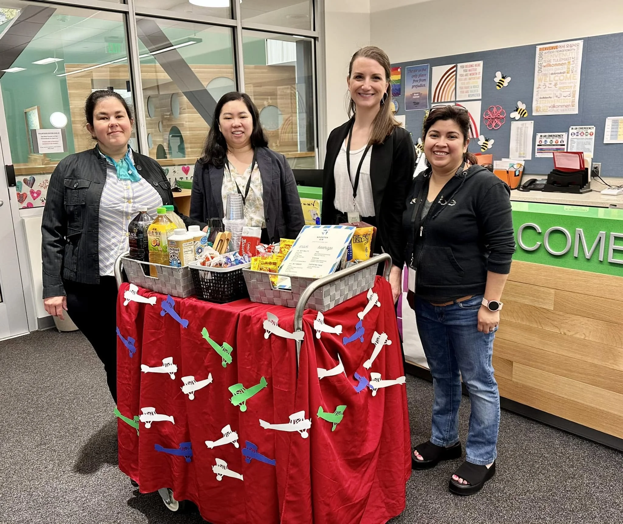 Four women standing behind a table with snacks and beverages in a room decorated with positive messages and rainbow flags, suggesting a community event or celebration.
