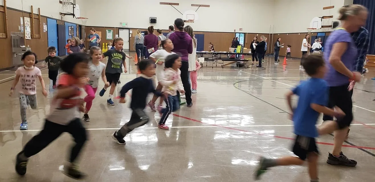 Children running and playing in a gymnasium with adults standing and talking in the background.