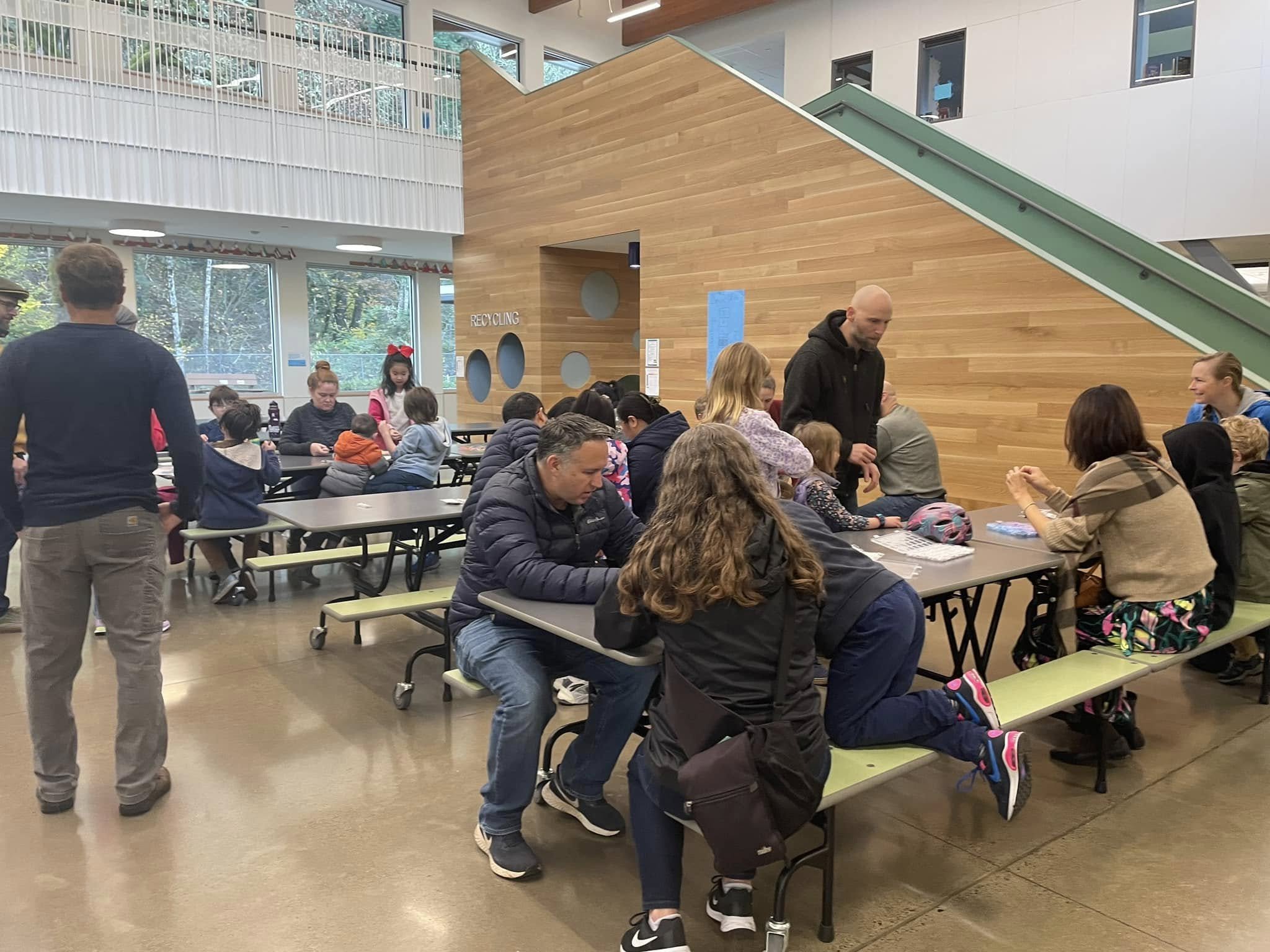 People gathered in a school or community cafeteria, sitting and standing around tables. A wooden wall with circular cutouts and a sign reading 'Recycling' are in the background. Large windows let in natural light, and a staircase is visible on the right side of the image.
