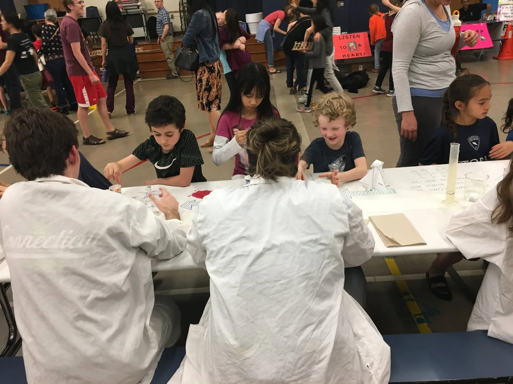 Children and adults gathered at a science fair table, with some children wearing protective lab coats, participating in a science experiment involving a test tube and water. People in the background are walking and engaging at various booths, with signs and displays visible.