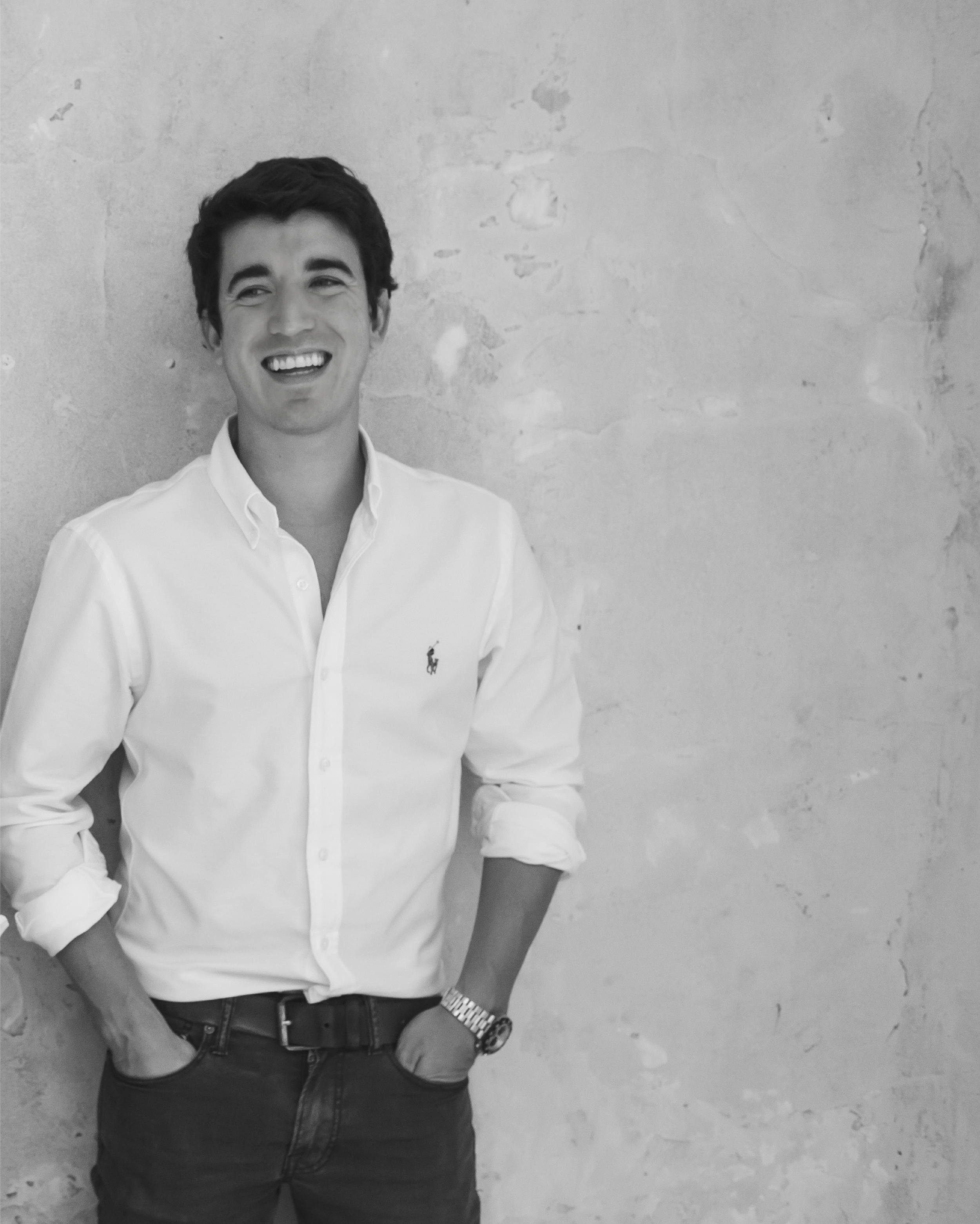 Black and white photo of a smiling young man with dark hair wearing a white button-up shirt and dark pants, standing against a textured wall with his hands in his pockets.