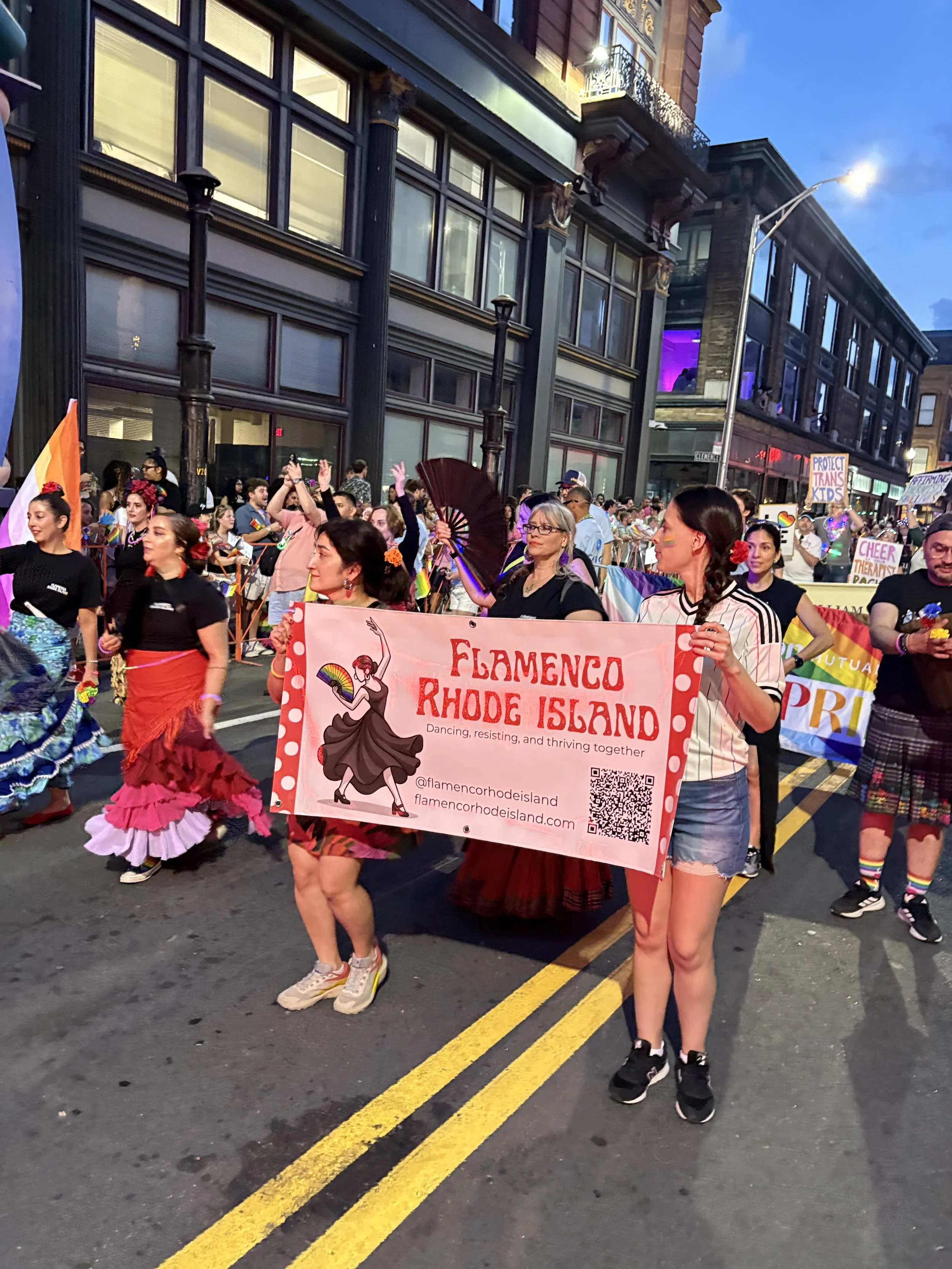 People marching in a pride parade, holding a pink banner that reads 'Flamenco Rhode Island' with a girl dancing with a fan illustration. The crowd includes individuals dressed in colorful and traditional flamenco attire, with some wearing rainbow accessories, on a city street with buildings and streetlights in the evening.