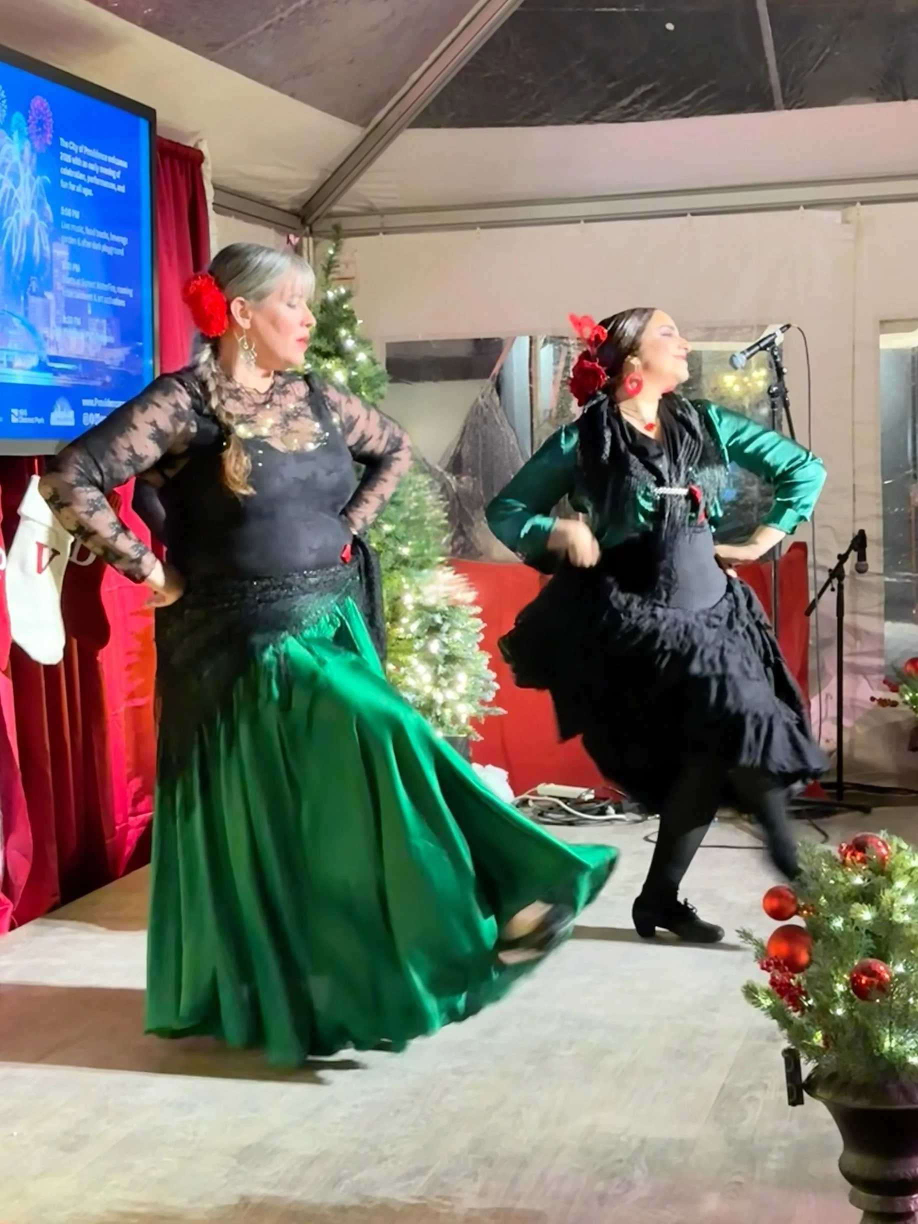 Two women dressed in traditional Mexican attire performing a folkloric dance on stage, with Christmas decorations including a small decorated tree and ornaments in the background, inside a festive setting.