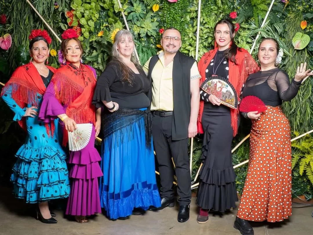 Group of six people dressed in traditional Spanish flamenco attire standing in front of a lush green tropical backdrop with flowers. They are posing for a photo, smiling and holding fans.