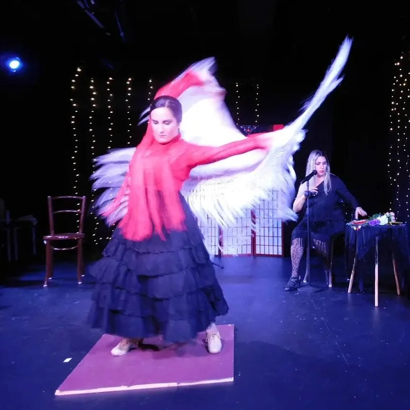 Performer in a flowing red and black dress dancing on stage with large white wings, stage is decorated with string lights, woman sits at a table with flowers and a microphone nearby.