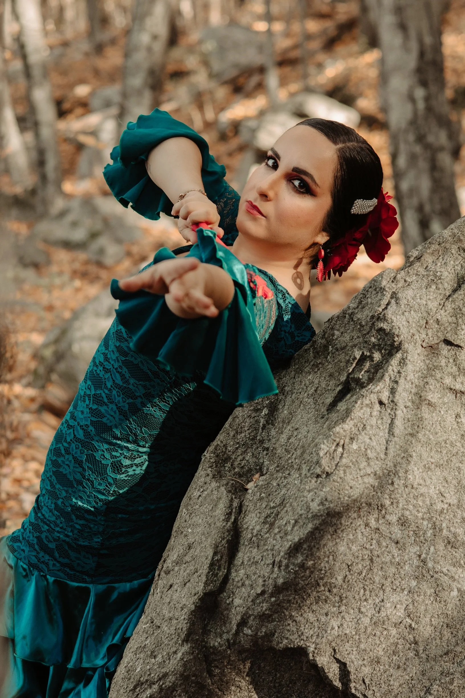 A woman dressed in traditional Mexican attire, with dark hair adorned with red flowers and a pearl hairpin, leaning against a large rock in a forest setting, reaching out with her hand.