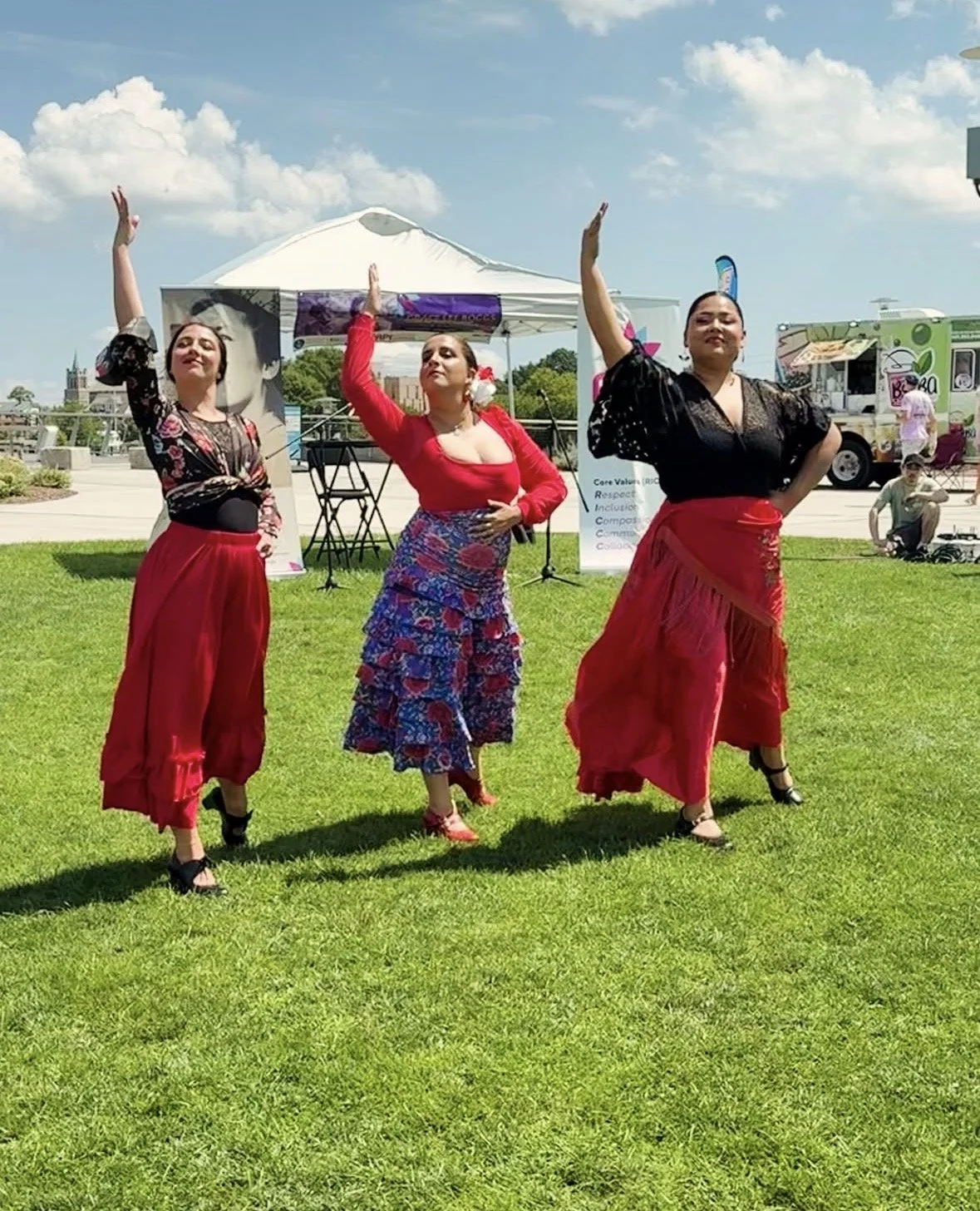 Three women dressed in colorful skirts and blouses performing a dance on a grassy outdoor area during a sunny day with cloudy sky, with tents, food trucks, and people in the background.