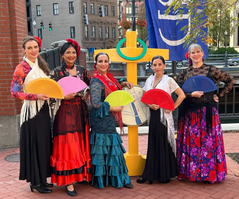 Five women dressed in traditional Spanish flamenco attire standing outdoors with colorful fans, in front of a large yellow cross sculpture and a blue flag, city buildings in the background.