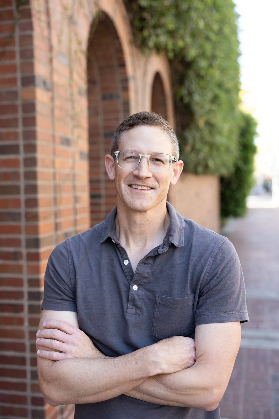 A smiling man with glasses standing outdoors with arms crossed, in front of a brick wall with arches and greenery.